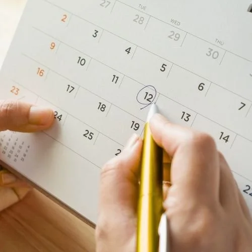 A desktop calendar with "12" circled. A hand holding a gold mechanical pencil circled the date.