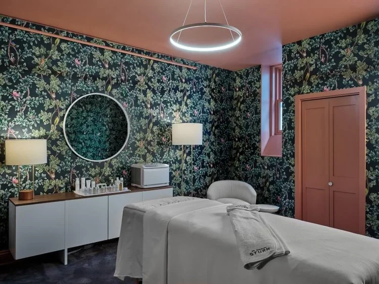 A stylish spa treatment room with floral wallpaper, a massage table covered in white linens, a round mirror, lamps, and a cabinet with skincare products. The ceiling is painted peach and features a modern circular light fixture.