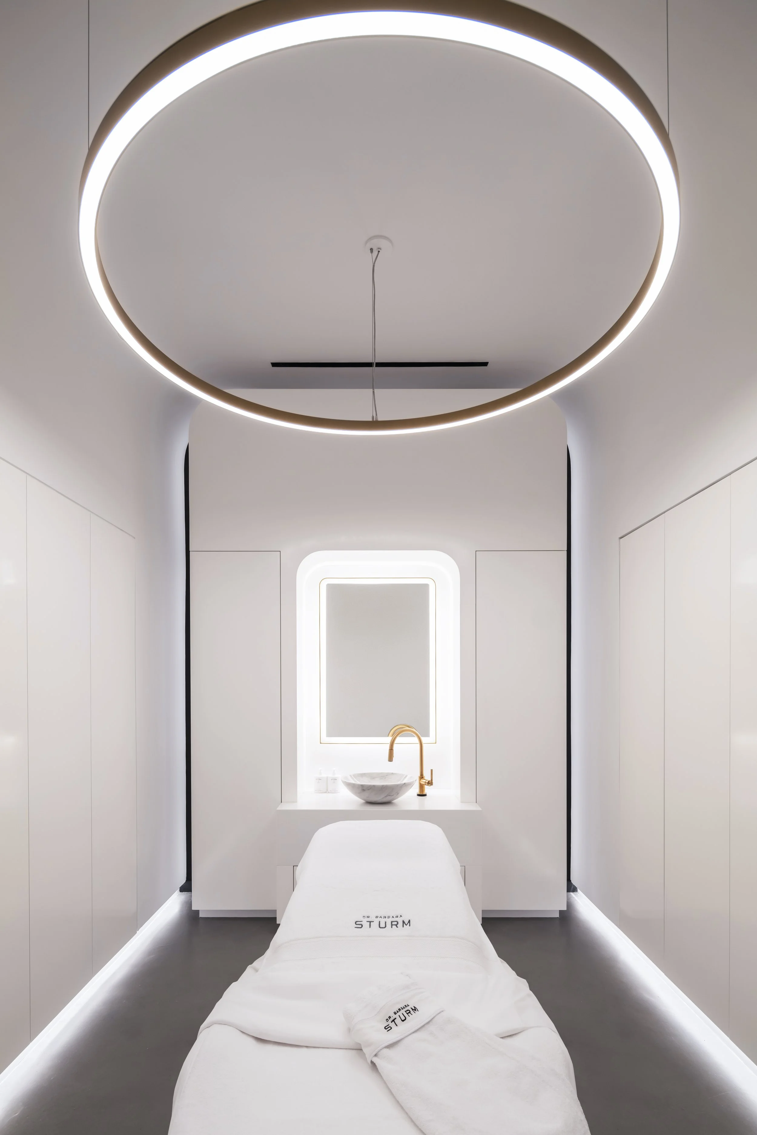 Minimalist spa room with white treatment table, circular ceiling light, and sink with mirror.