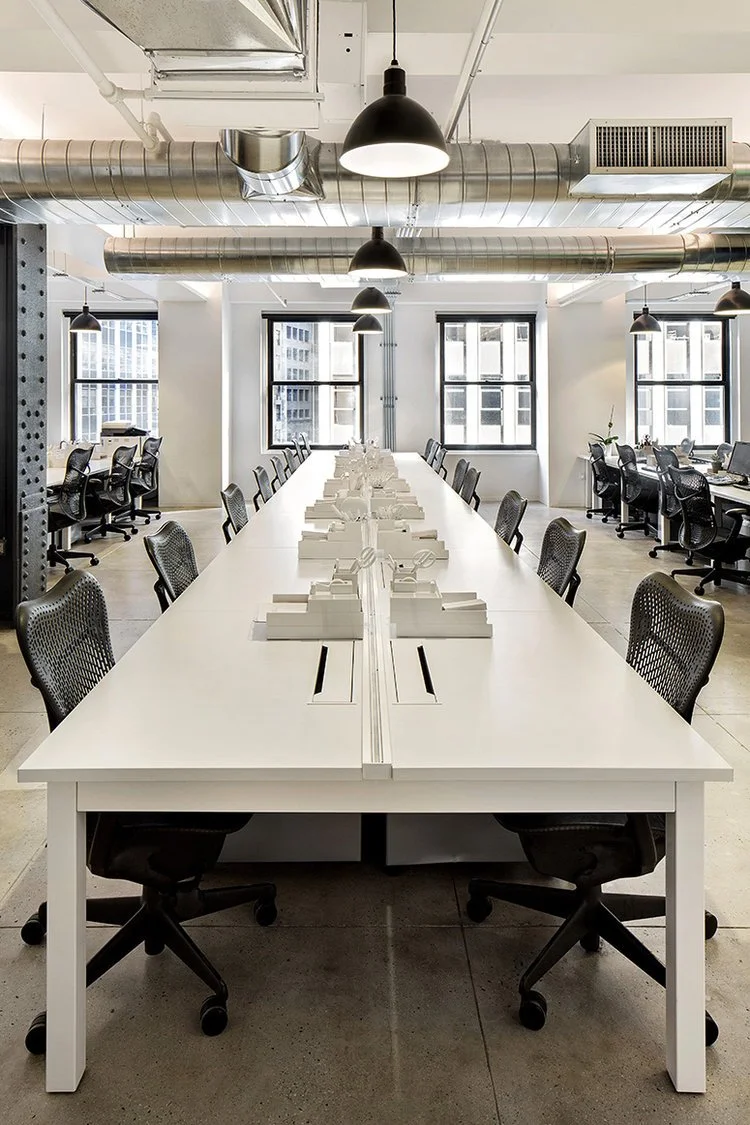 Modern office space with long white desks, black office chairs, and industrial-style light fixtures. Exposed ductwork and large windows enhance the open and airy atmosphere.