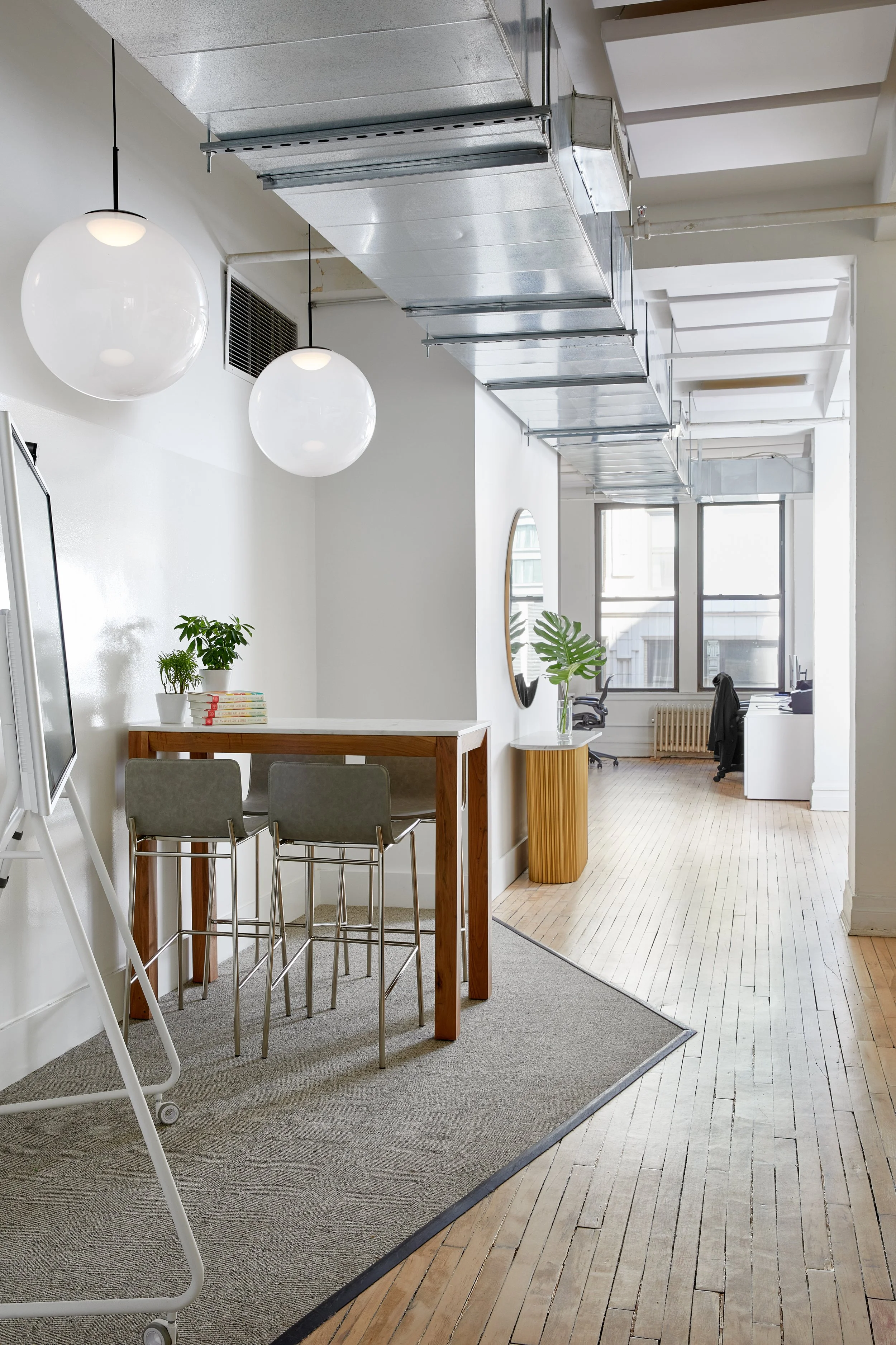 Modern office interior with a tall wooden table, chairs, hanging round lights, indoor plants, a wall mirror, wooden flooring, and exposed ductwork.