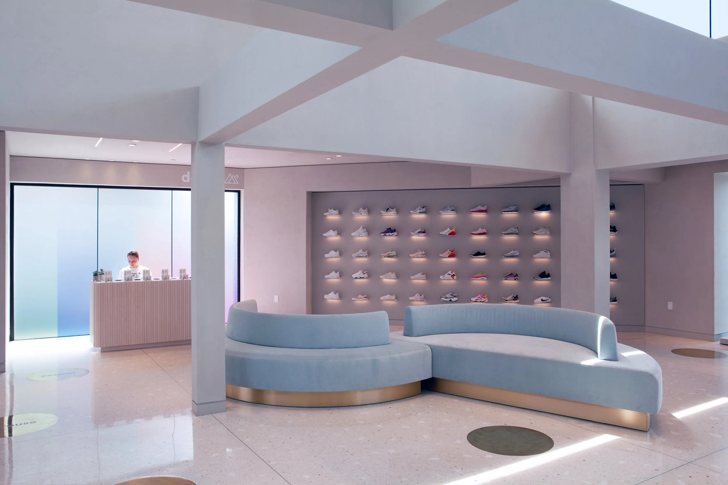 Modern shoe store interior with light blue seating and a wall display of sneakers. A reception desk is positioned under a large window with gradient colors. Bright and minimalistic design.