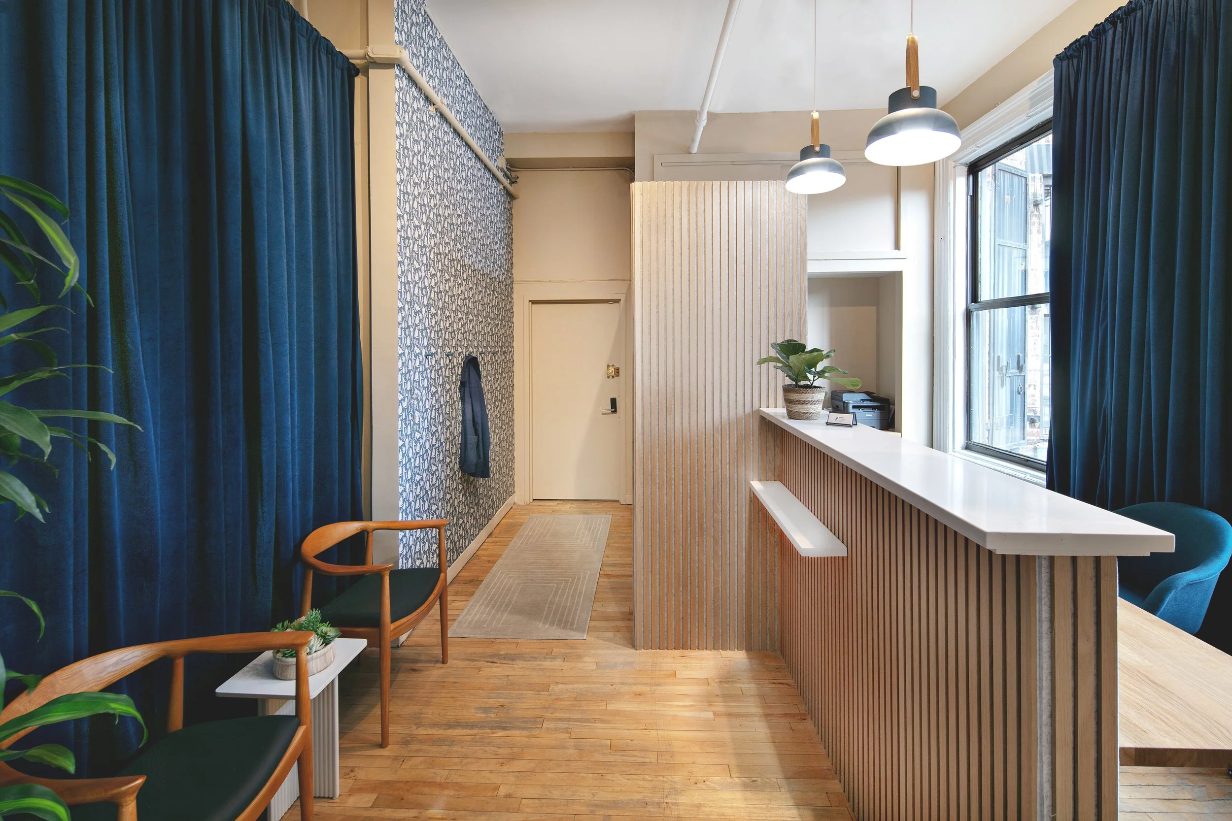 Modern office reception area with wooden flooring, blue curtains, a white counter, potted plant, and hanging lights.
