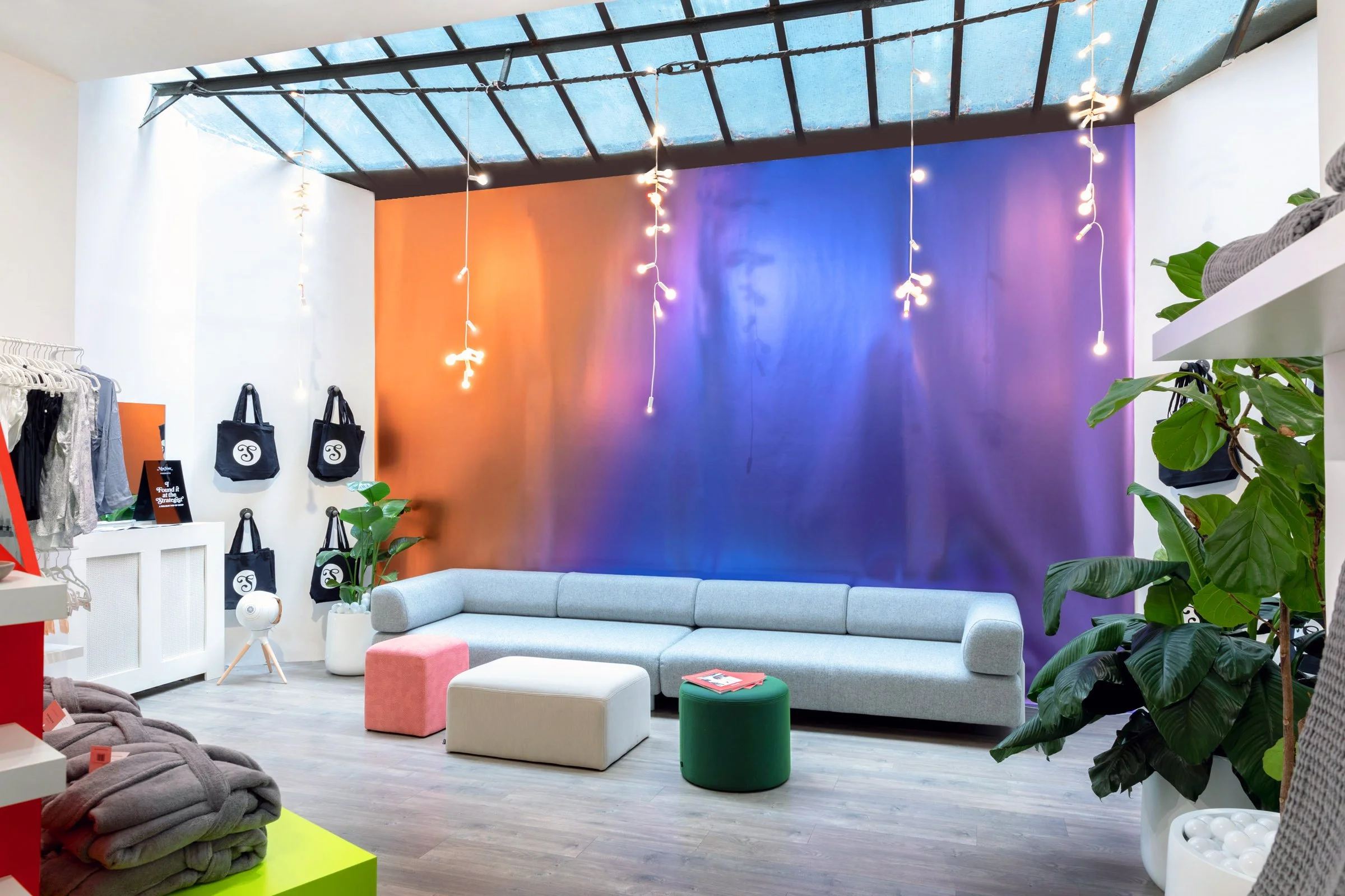 Modern interior with colorful gradient wall, gray sofa, hanging lights, plants, and shopping bags on display.