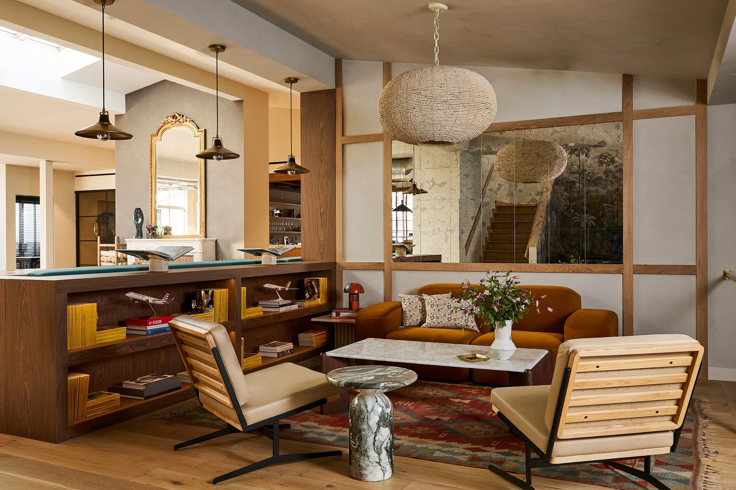 Living room with orange sofa, two beige chairs, marble coffee table, wooden bookshelf with model airplanes, decorative mirror, large woven pendant light, flowers on table, and wooden staircase in background.