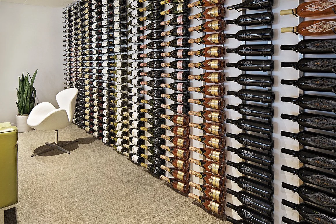 Wall-mounted wine bottle display with various bottles, modern white chair, and a potted plant on carpeted floor.