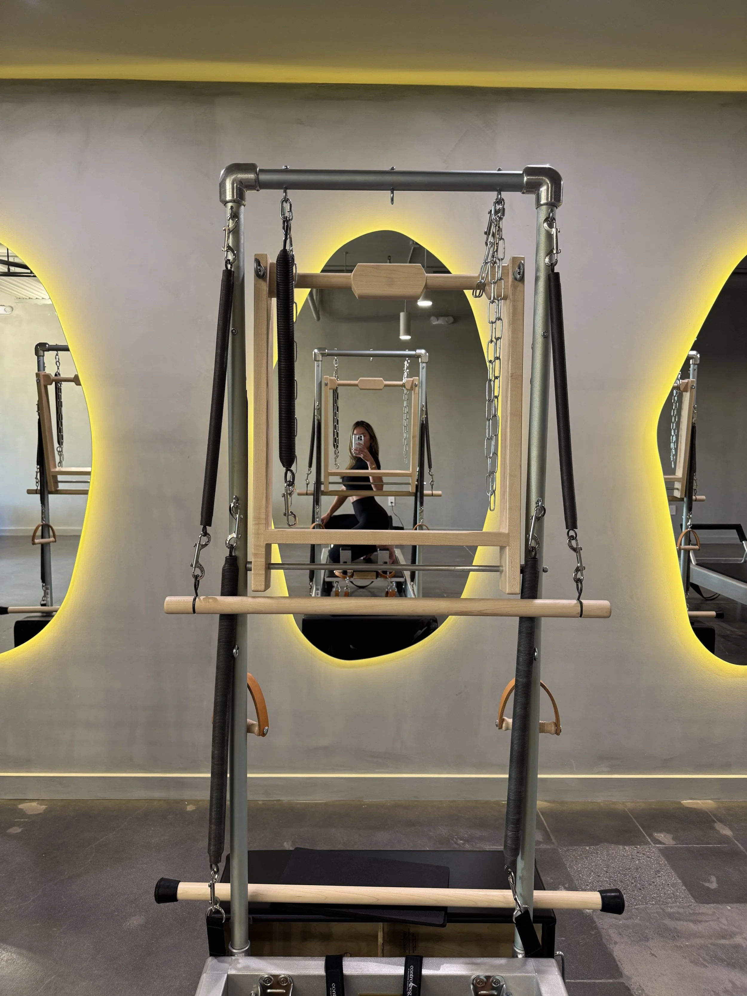 Pilates exercise equipment with mirrors and a person taking a photo in the reflection. The room has a modern design with yellow backlighting.