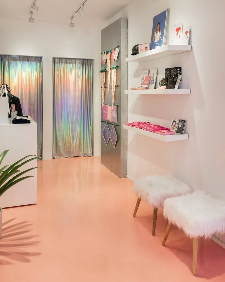 Boutique interior with pink floor, white walls, and shelves displaying merchandise. Two furry stools and a plant are visible. Iridescent curtains in the background add a colorful touch.
