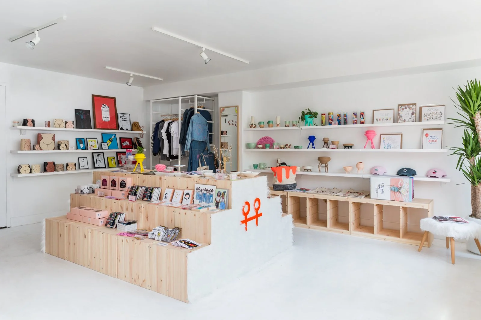 Interior of a modern retail store with shelves displaying artwork, home decor, clothing, and accessories. Wooden displays hold books, framed art, and unique crafts. Clothing racks feature jackets and shirts. A bright, minimalist design dominates the space.