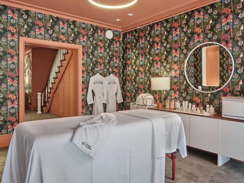 A plush spa treatment room with a bed covered in white linens labeled 'Sturm'. The room has vibrant floral wallpaper and a coral-colored ceiling. There are two white robes hanging on the wall. A circular mirror, lamp, and various skincare products are displayed on a countertop. Stairs are visible through an open doorway.