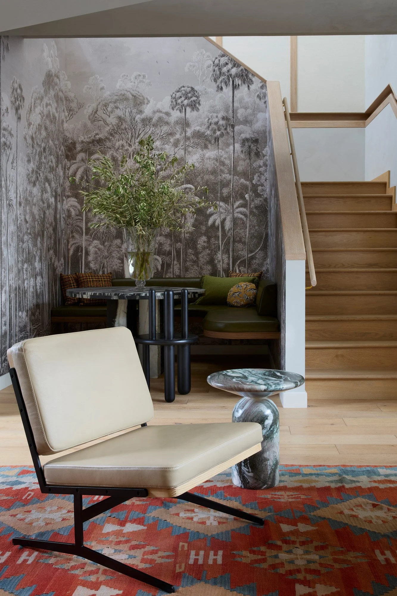 Modern living room with beige lounge chair, red patterned rug, marble side table, green bench with pillows, large glass vase with greenery, forest-themed wallpaper, wooden staircase.