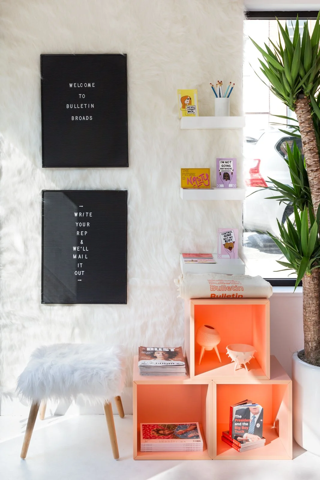 Room with white furry walls, featuring a stool with a similar fur seat, orange cubes with magazines and planters, and two felt boards with text. Shelves hold books, a pencil holder, and a plant is visible by the window.