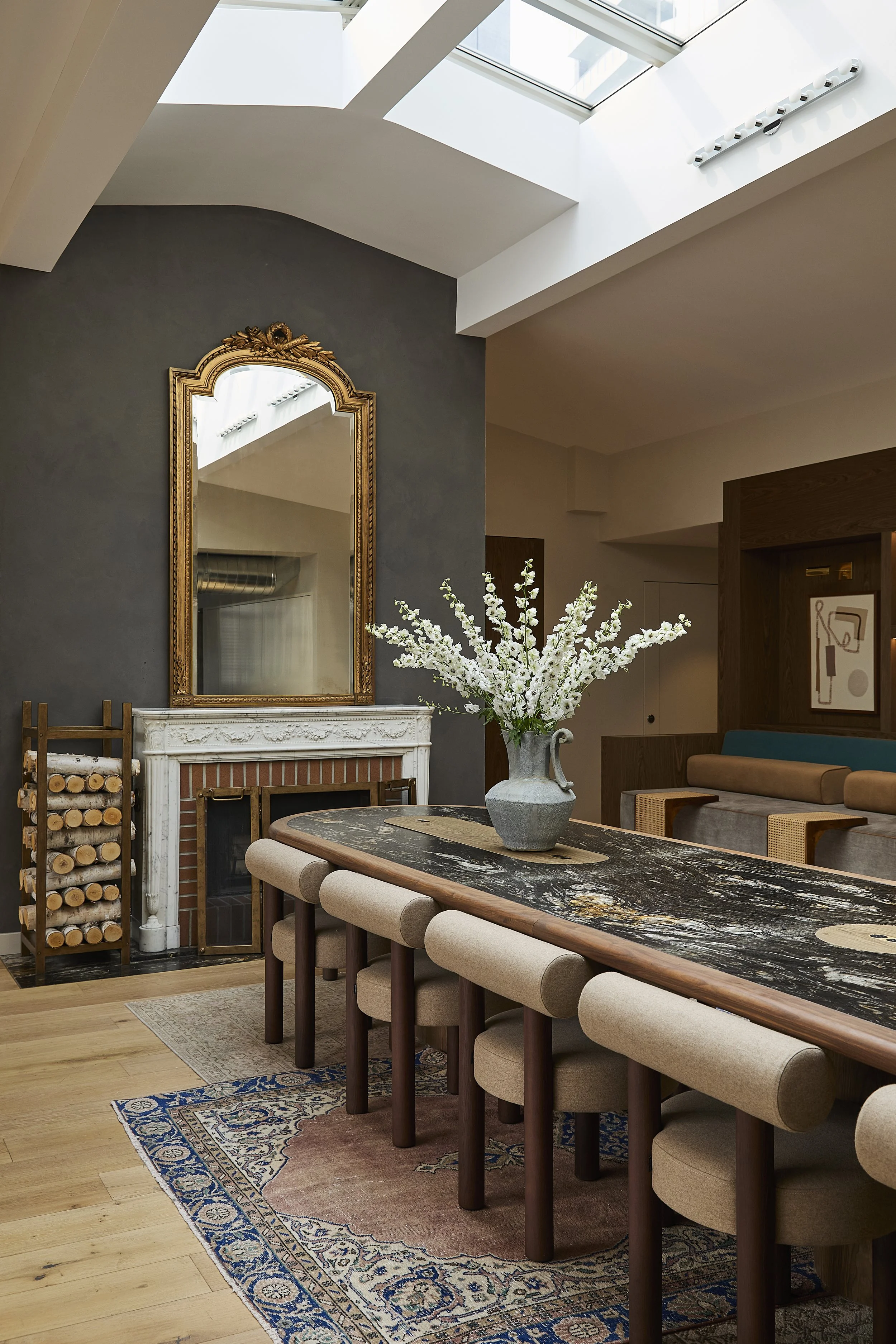 Interior of a dining room with a large marble-topped table, beige upholstered chairs, a decorative fireplace with a gold mirror above it, a vase of white flowers on the table, and a skylight overhead.