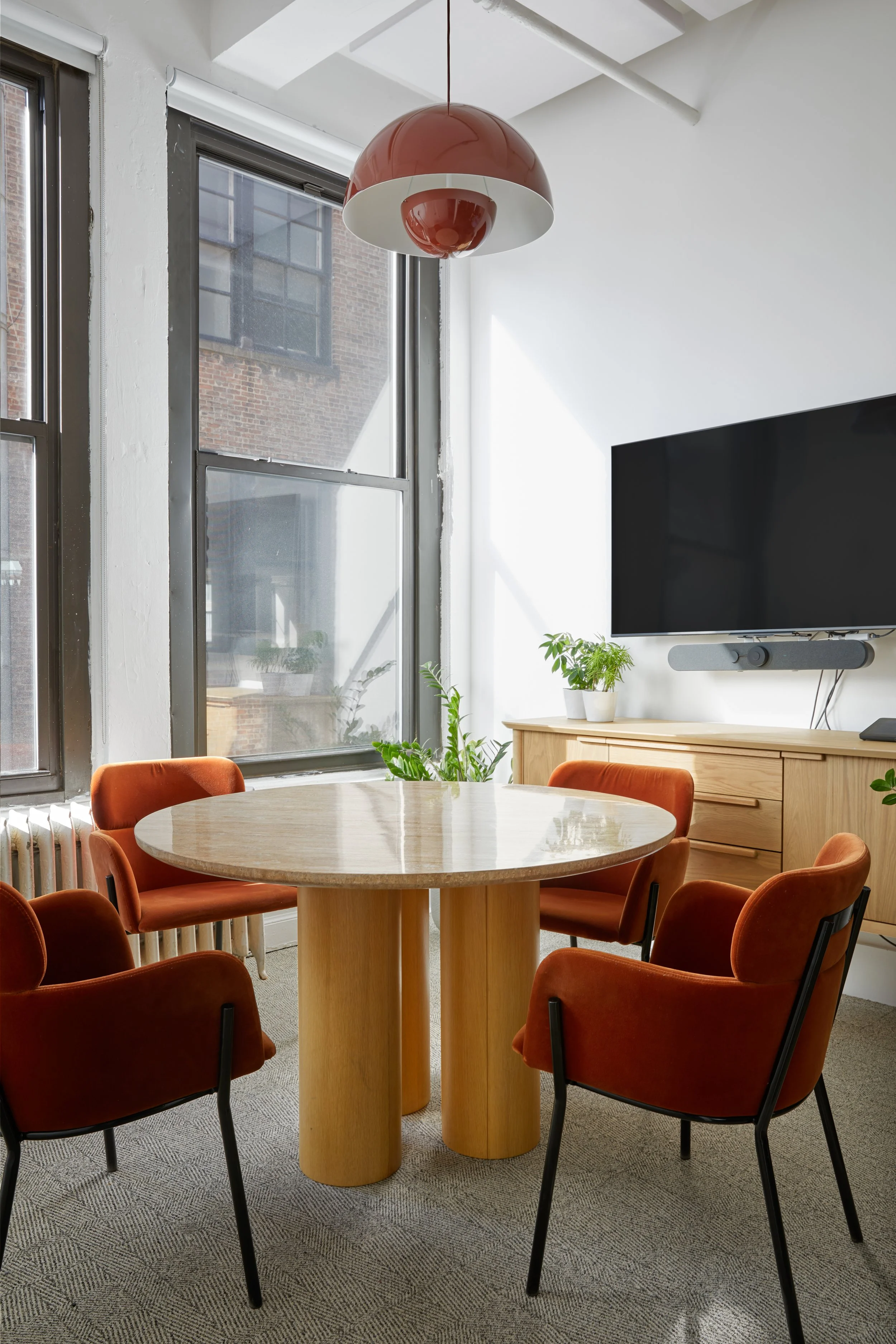 Modern office space with a round wooden table, orange chairs, a flat screen TV, and large windows with natural light.