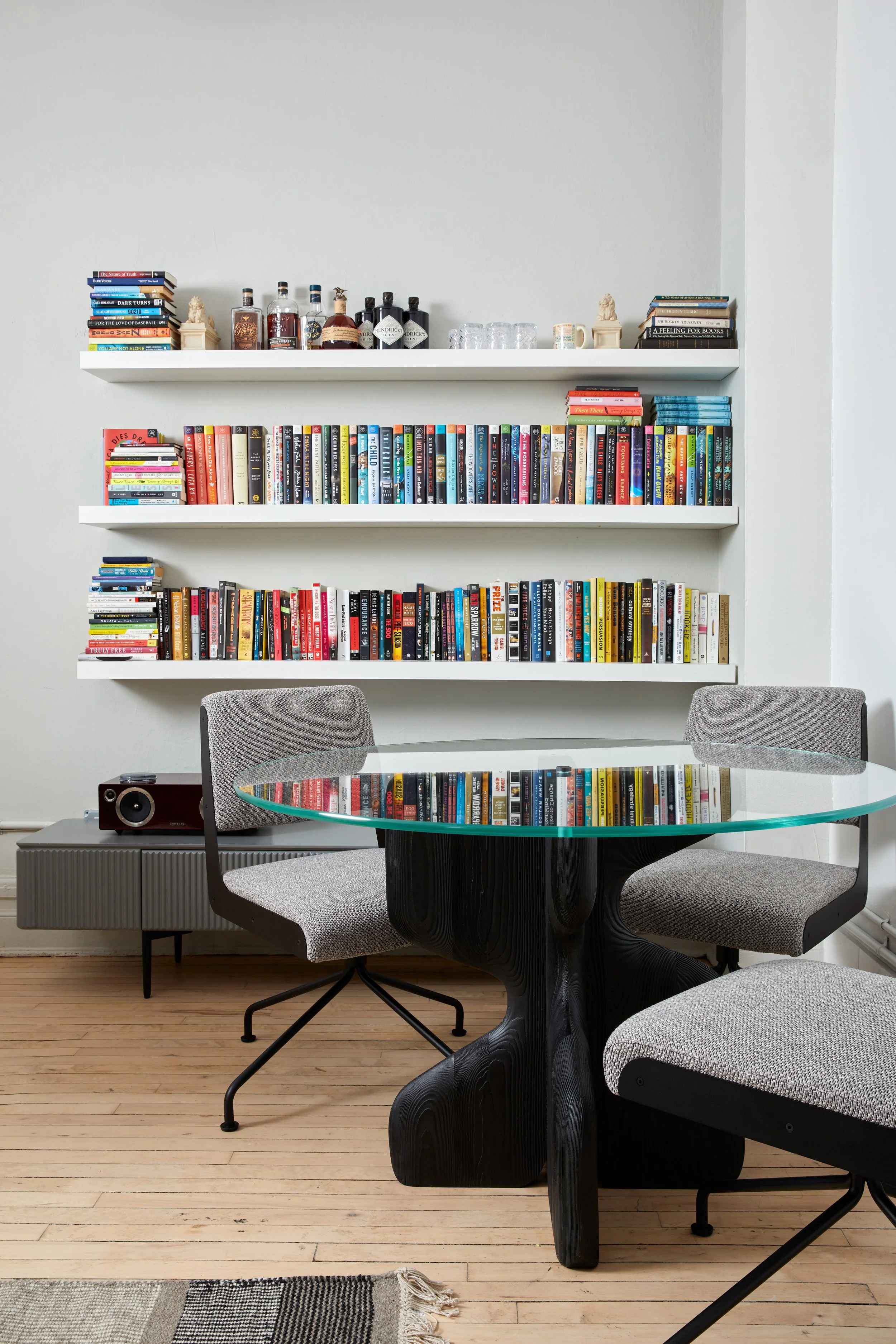 Modern living room with a glass round table and gray chairs on a wooden floor, featuring two wall-mounted bookshelves filled with various books, decorative items, liquor bottles, and glasses.