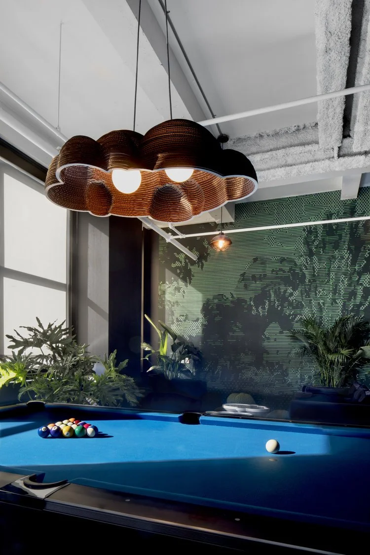Contemporary room with a blue pool table and a unique cloud-shaped pendant light. The room features large windows, indoor plants, and a textured wall with green accents.