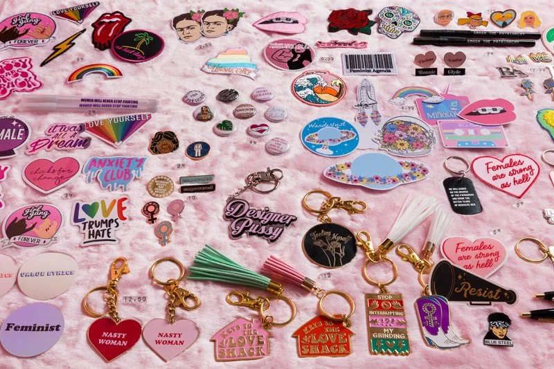 A collection of colorful pins, patches, keychains, and stickers with various designs and slogans on a pink background.