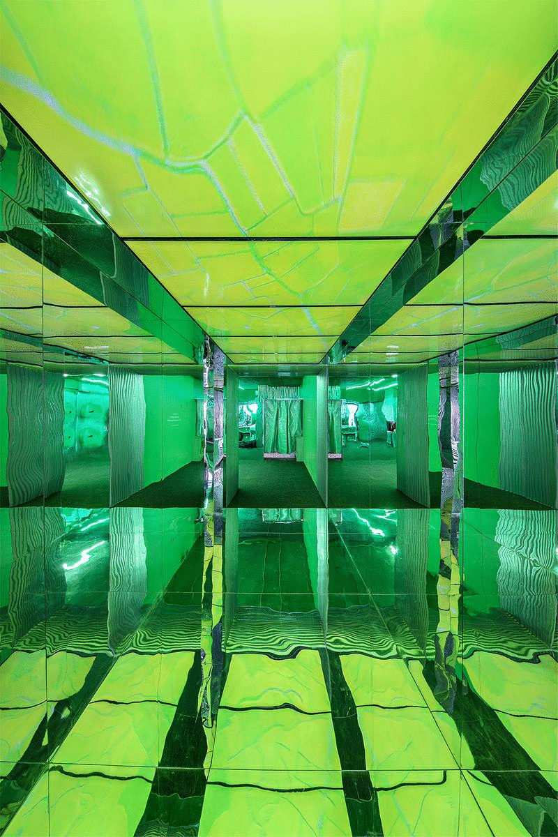 Interior view of a room with reflective surfaces and green lighting, creating an optical illusion effect. The walls, floor, and ceiling are covered in mirrors, with distorted reflections of lines and patterns, giving a futuristic and surreal appearance.