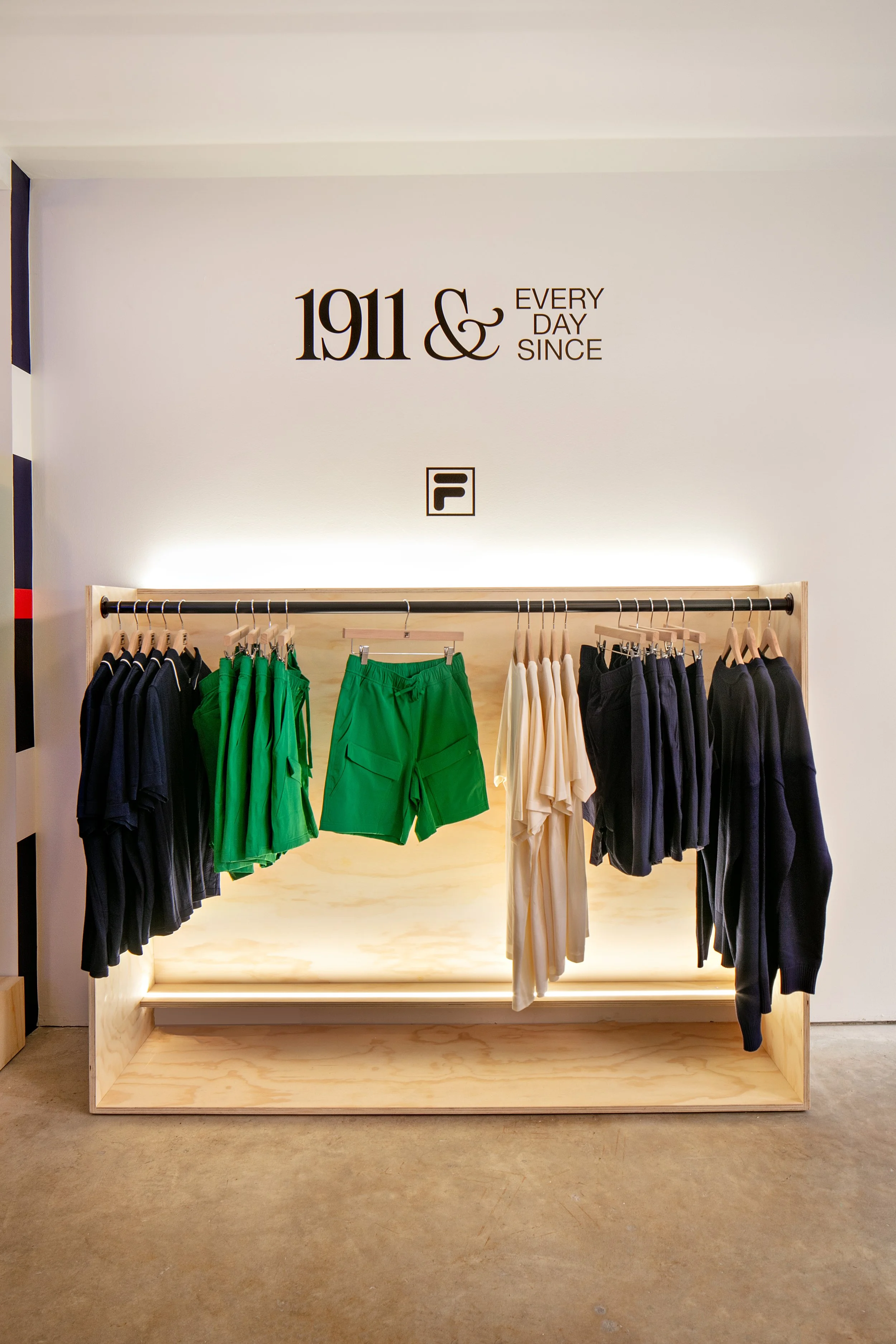 Clothing display in a store, featuring apparel on hangers, including green shorts and a mix of dark and light shirts. The background wall reads '1911 & Every Day Since', with a logo below.