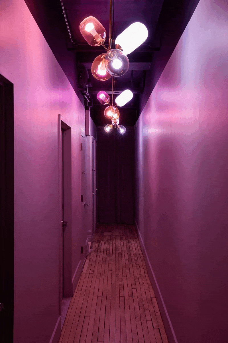 Narrow hallway with pink and purple lighting, wooden floor, and decorative ceiling light fixtures.