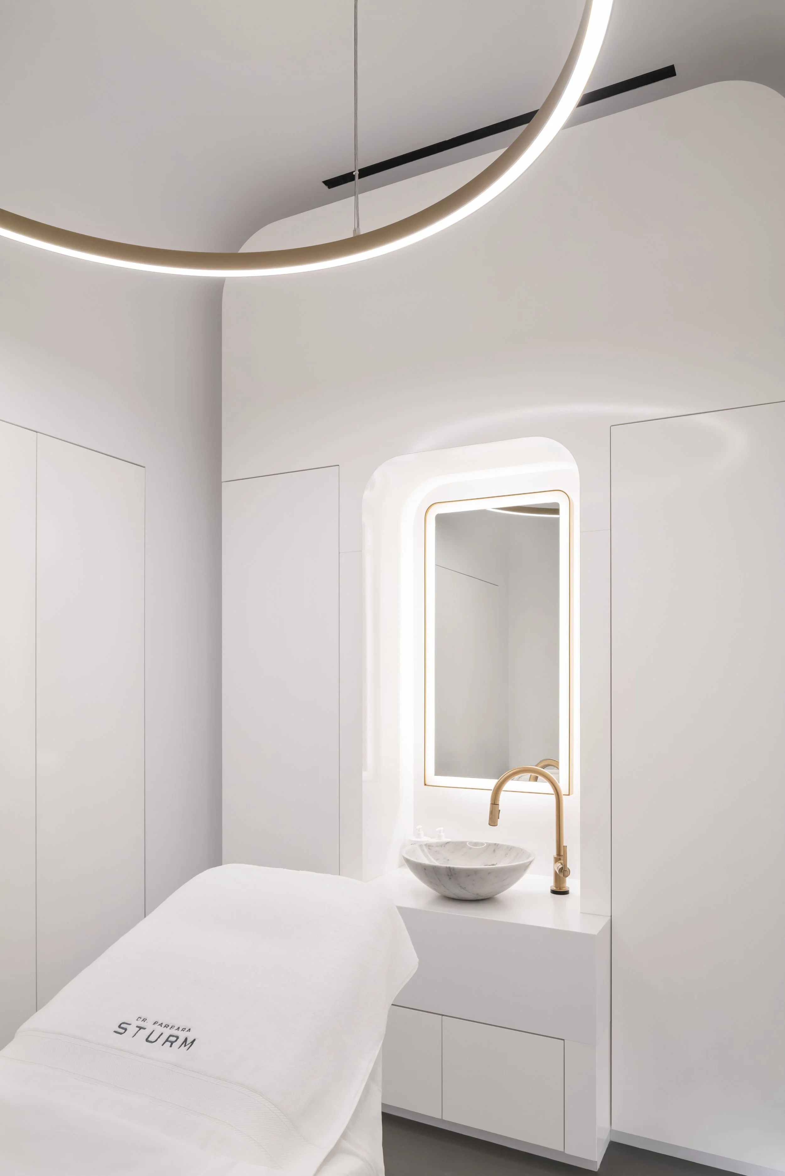 Modern minimalist spa room with white decor, circular overhead light, sink with gold faucet, and treatment table covered in white towel with "Dr. Barbara Sturm" text.