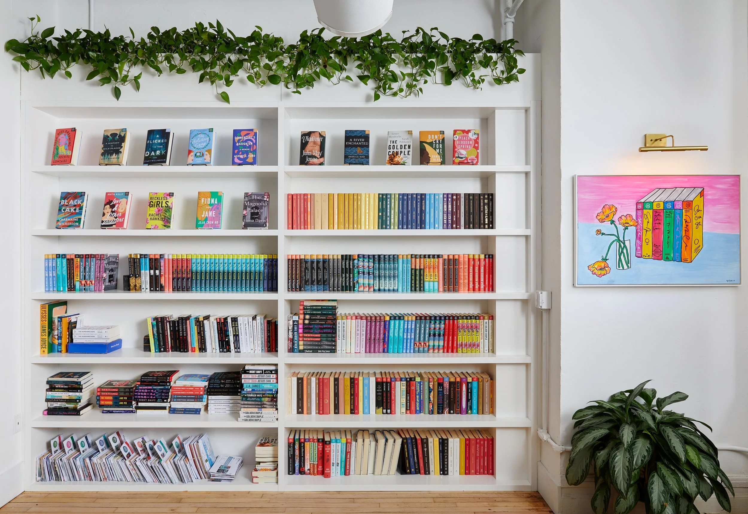 Bookshelf with colorful books, a wall art piece of books and flowers, green plant, and wooden floor.