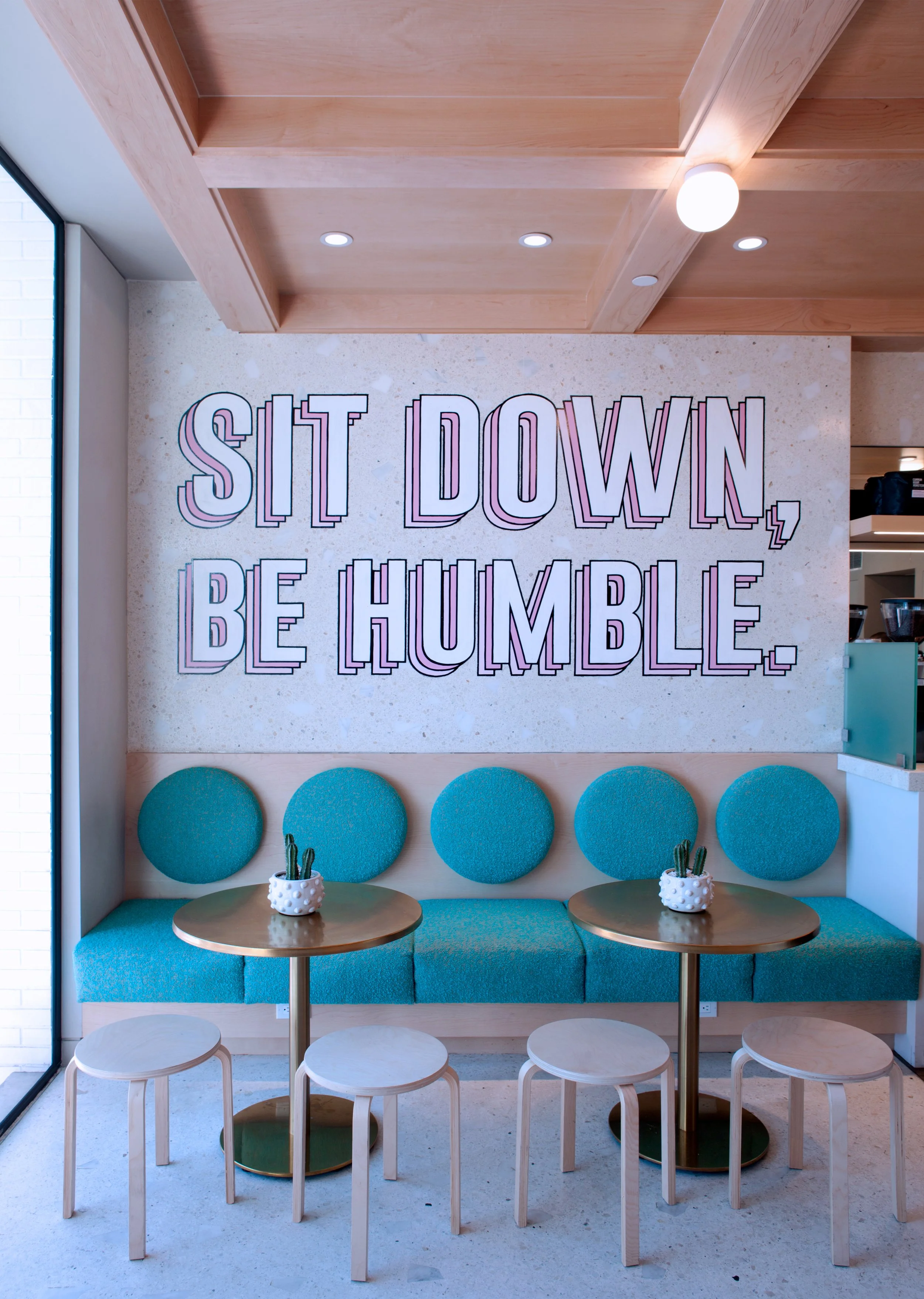 Café interior with minimalist design, featuring a wall with the text "Sit Down, Be Humble." Circular blue cushions line the bench seating, and small round tables with white stools and potted plants add to the décor.