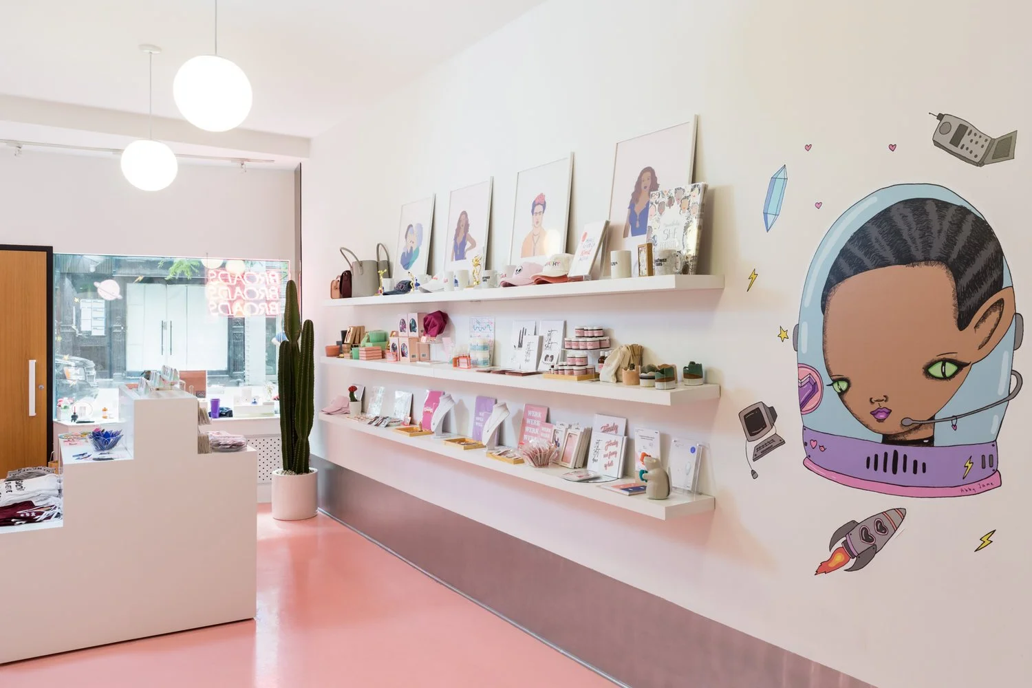 Boutique store interior with shelves displaying various gift items, framed art, and accessories like bags and hats. A mural featuring a cartoon character in a space helmet decorates the wall. A cactus is placed near the checkout counter.