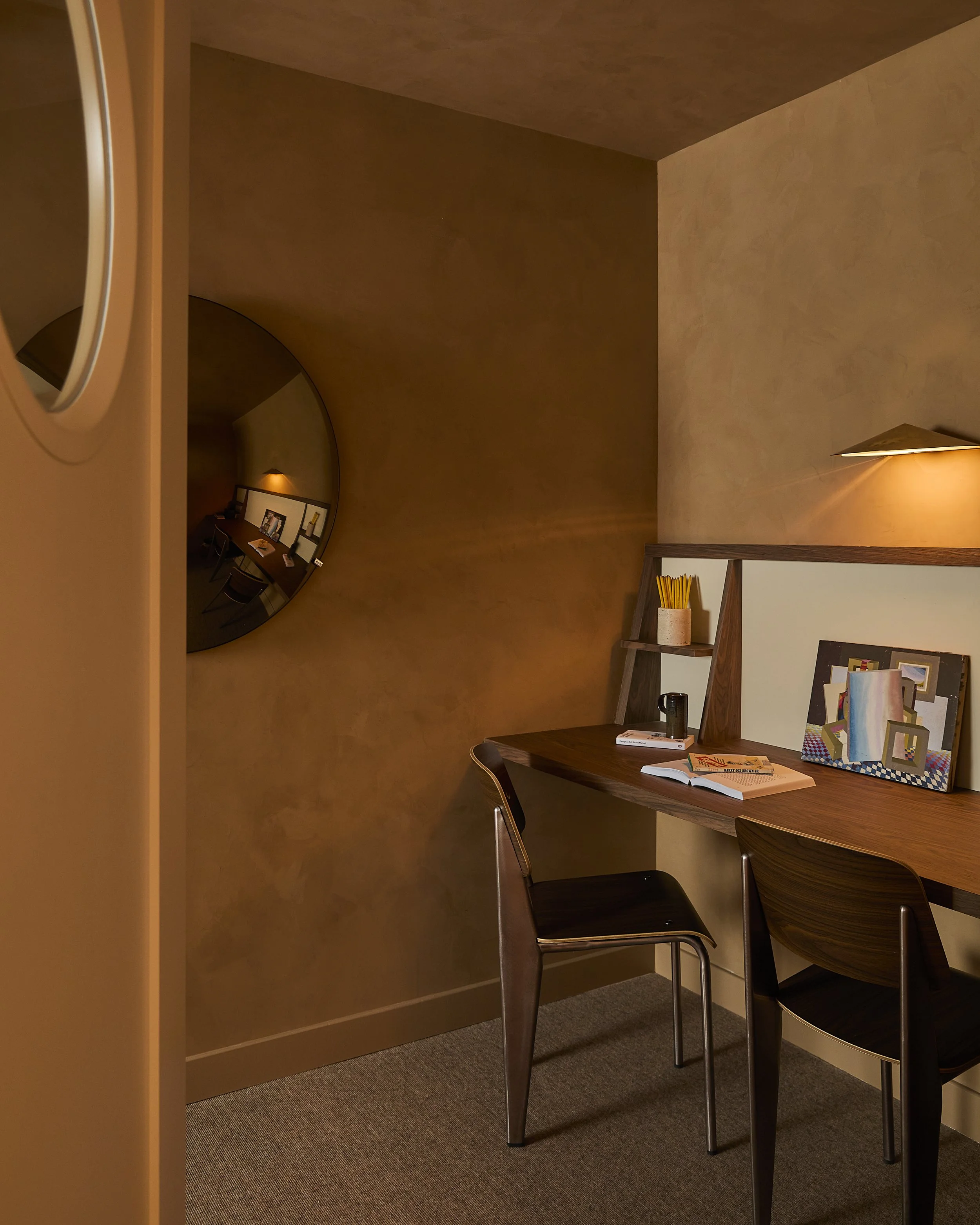 A cozy workspace with a wooden desk, two chairs, a small shelf with pencils, and framed artwork. A large circular mirror reflects part of the room.