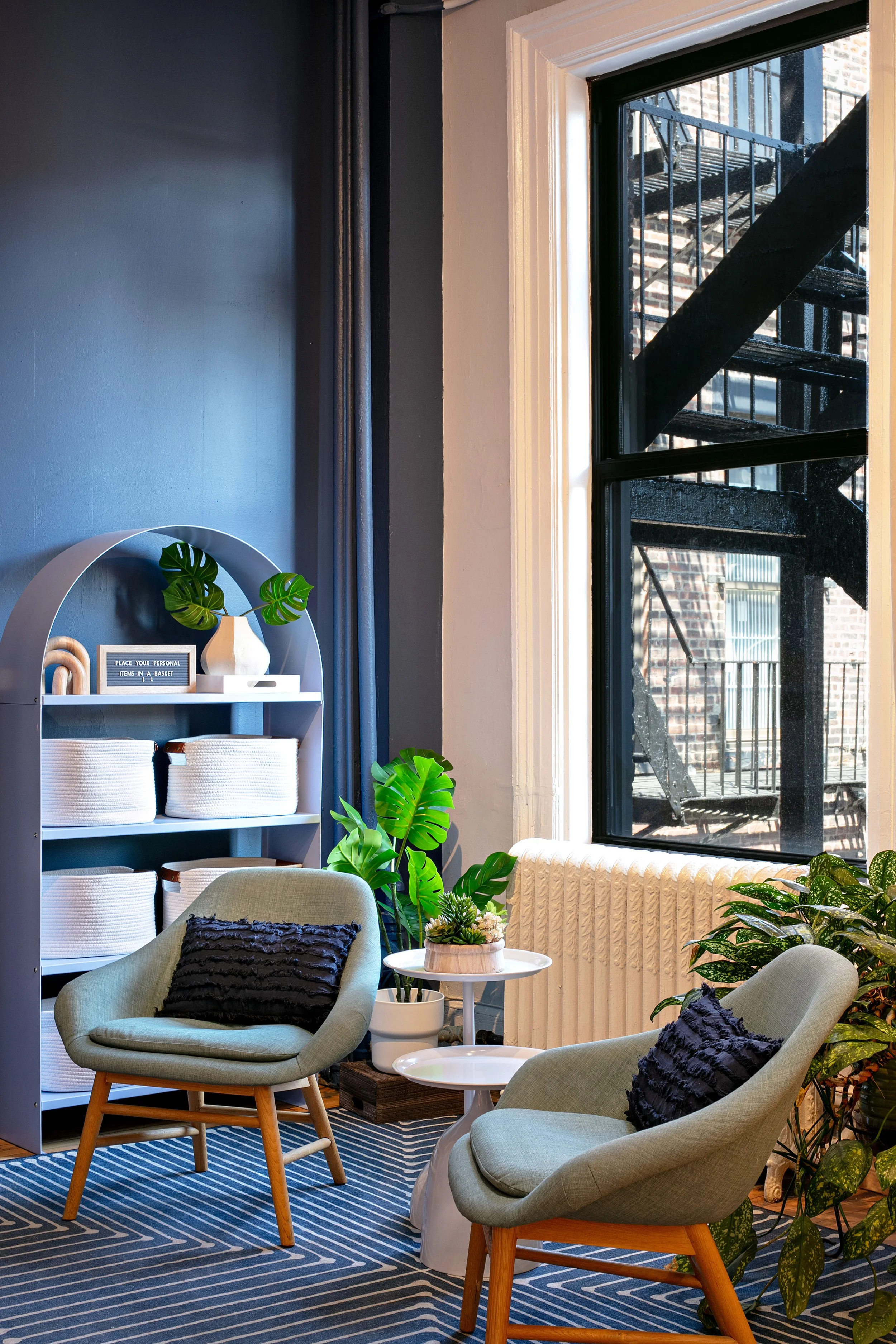 A cozy living room corner with two green armchairs and black accent pillows, a small white side table, and various potted plants. A gray bookshelf holds baskets and decor items. A window shows a view of an external staircase. The room features a patterned blue and white rug and blue painted walls.