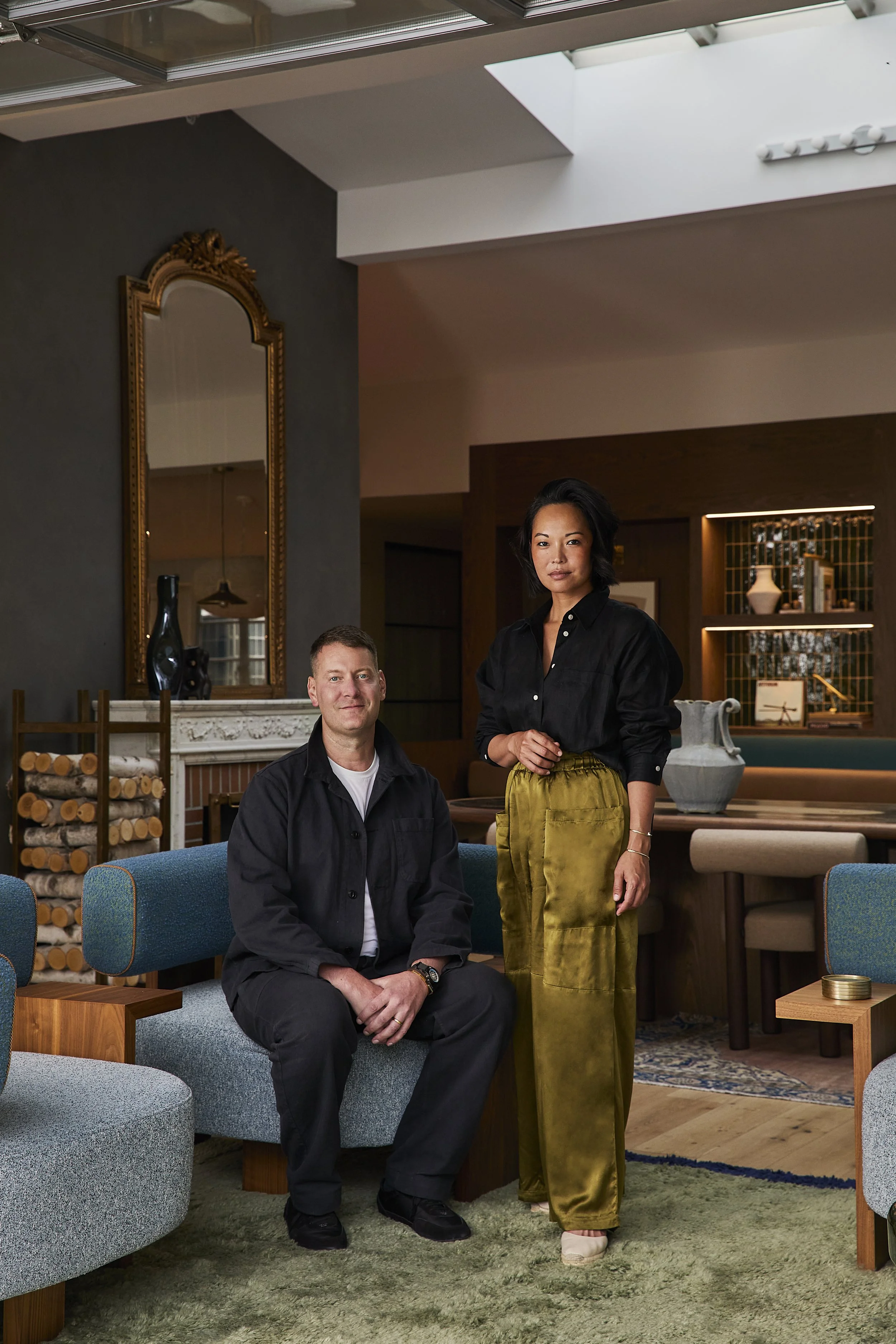 A man and woman in stylish clothing in a living room with modern decor, sitting and standing respectively, near a sofa and an artful bookshelf.