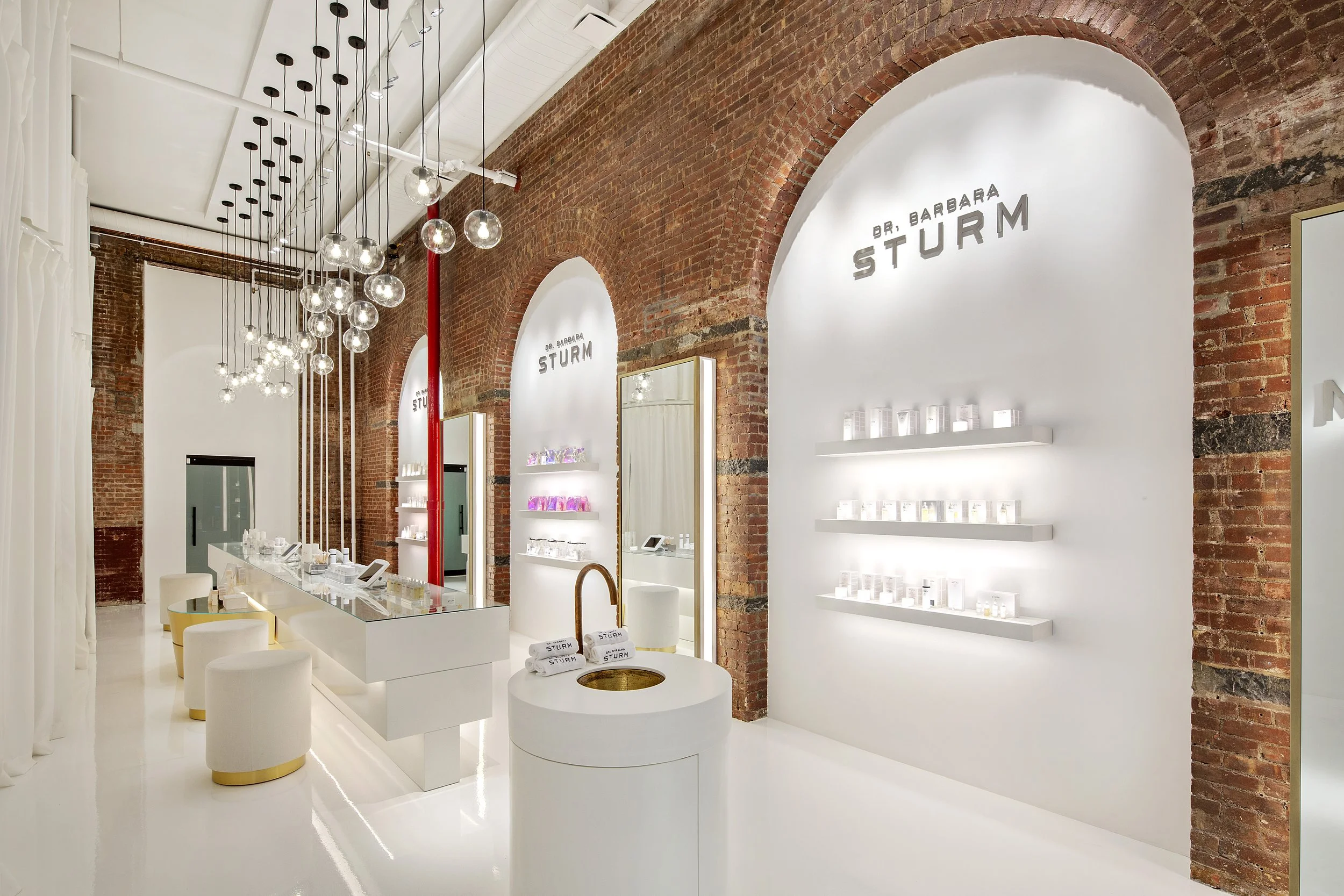 Modern skincare store interior with white decor, exposed brick walls, and a central table with products. Shelves display products on arched alcoves. Several spherical pendant lights hang from the ceiling.