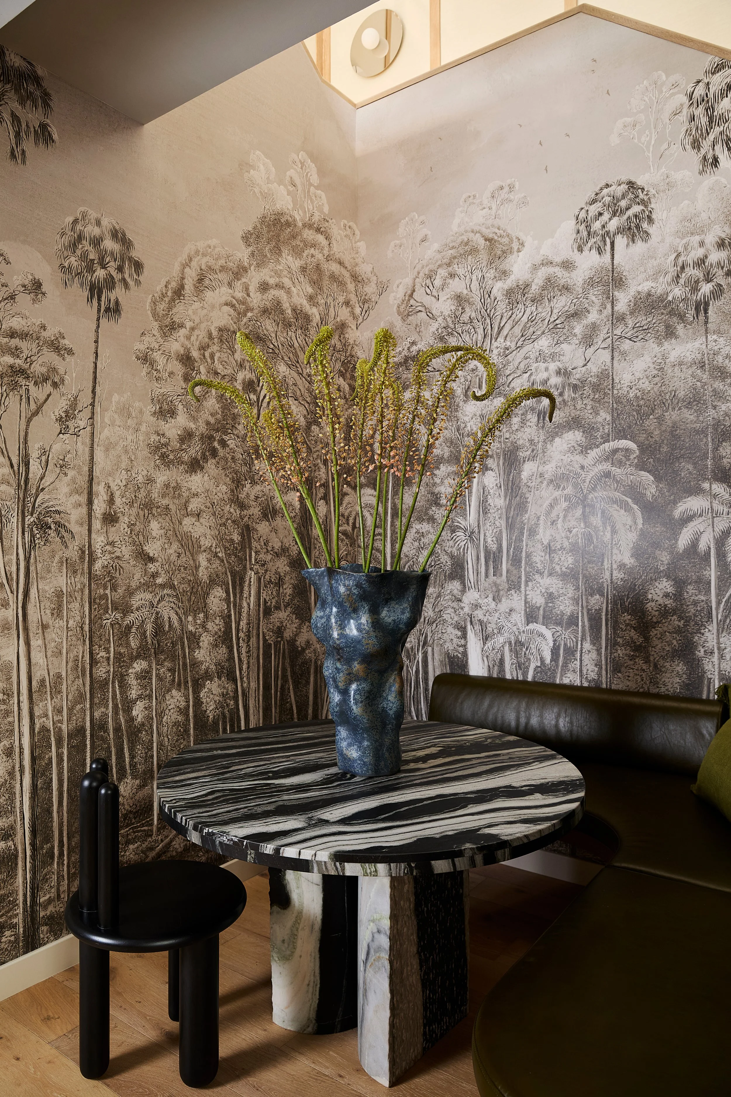 A round black and white marble table with a large textured blue vase holding tall, curly green and pink flowers, set against a wallpapered wall with a monochrome forest scene, in a modern interior with wooden flooring.