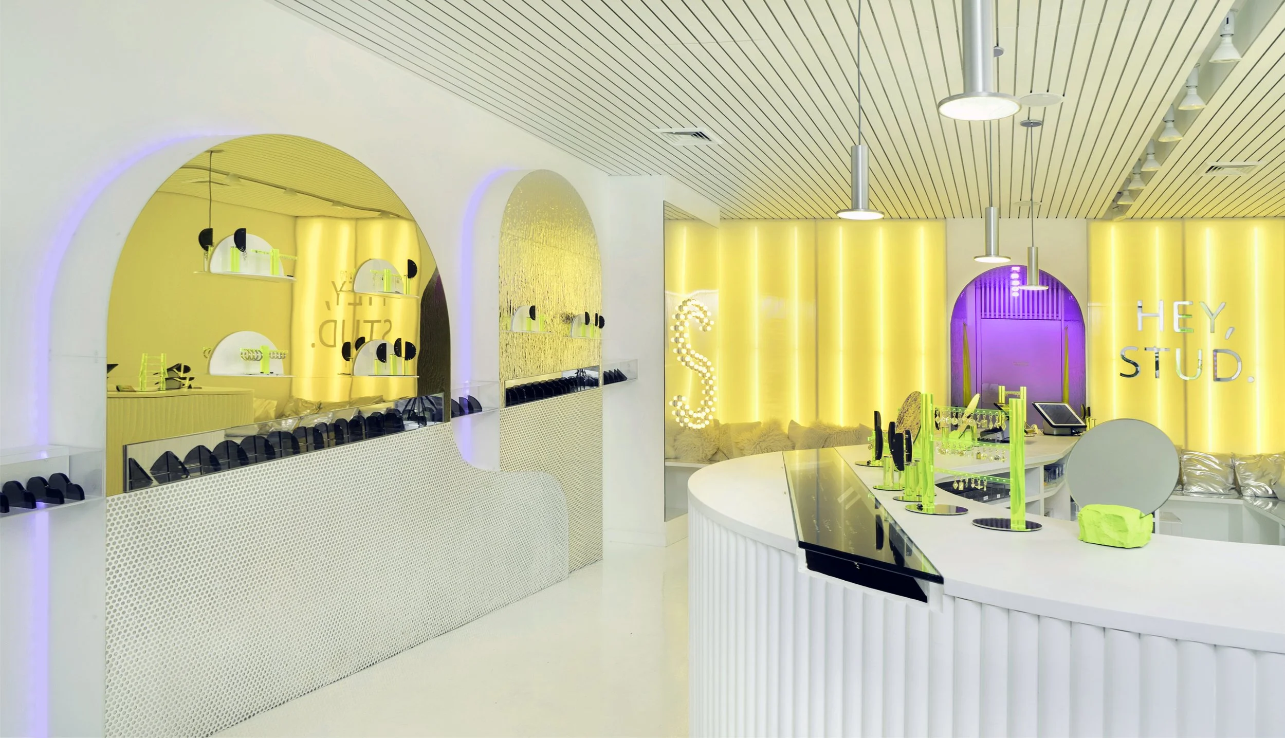 Modern retail store interior with neon accents, featuring display shelves, silver cushions, yellow and purple lighting, and "HEY, STUD." on a wall.