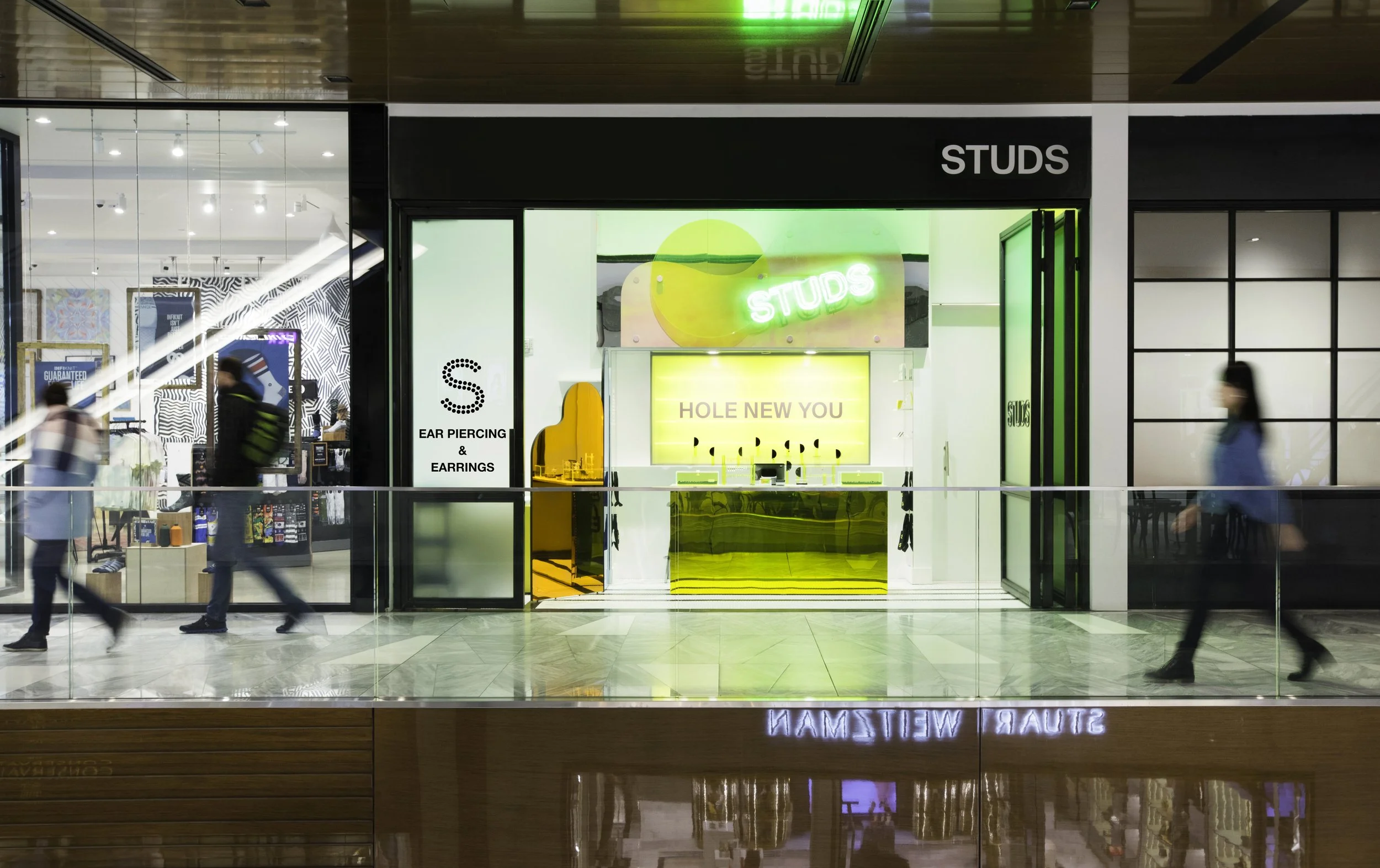 Storefront of Studs, an ear piercing and earrings shop, with people walking by. The interior has neon signs and modern decor.