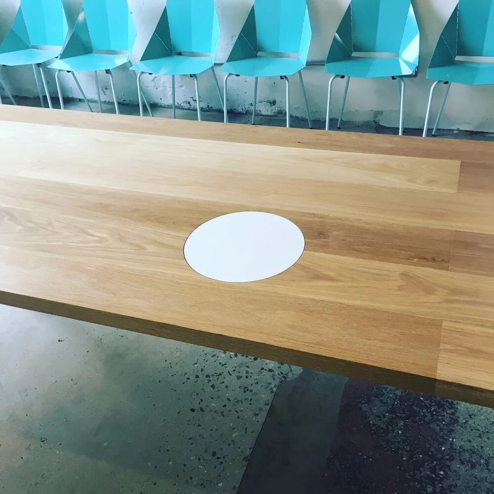 Wooden table with a white circular section and blue chairs in the background.