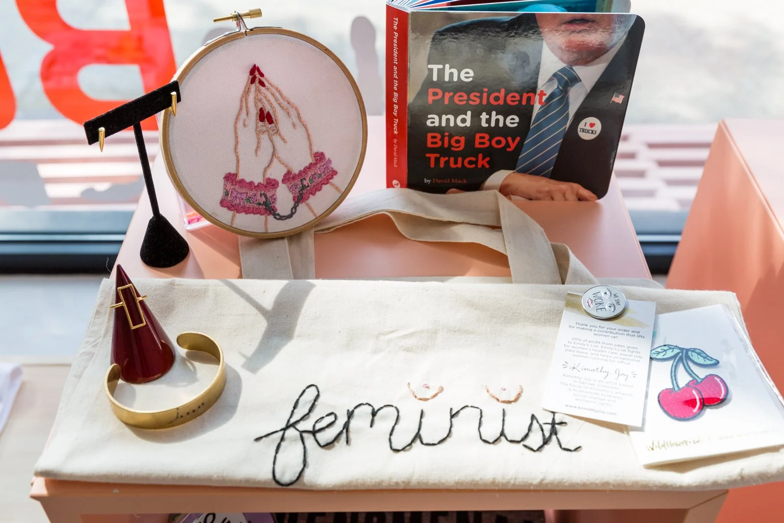 Tabletop display featuring a canvas tote bag with the word 'feminist' stitched on it, a book titled 'The President and the Big Boy Truck,' a hoop with embroidery of praying hands, a jewelry display with earrings, a ring on a stand, and some informational cards and a cherry patch.