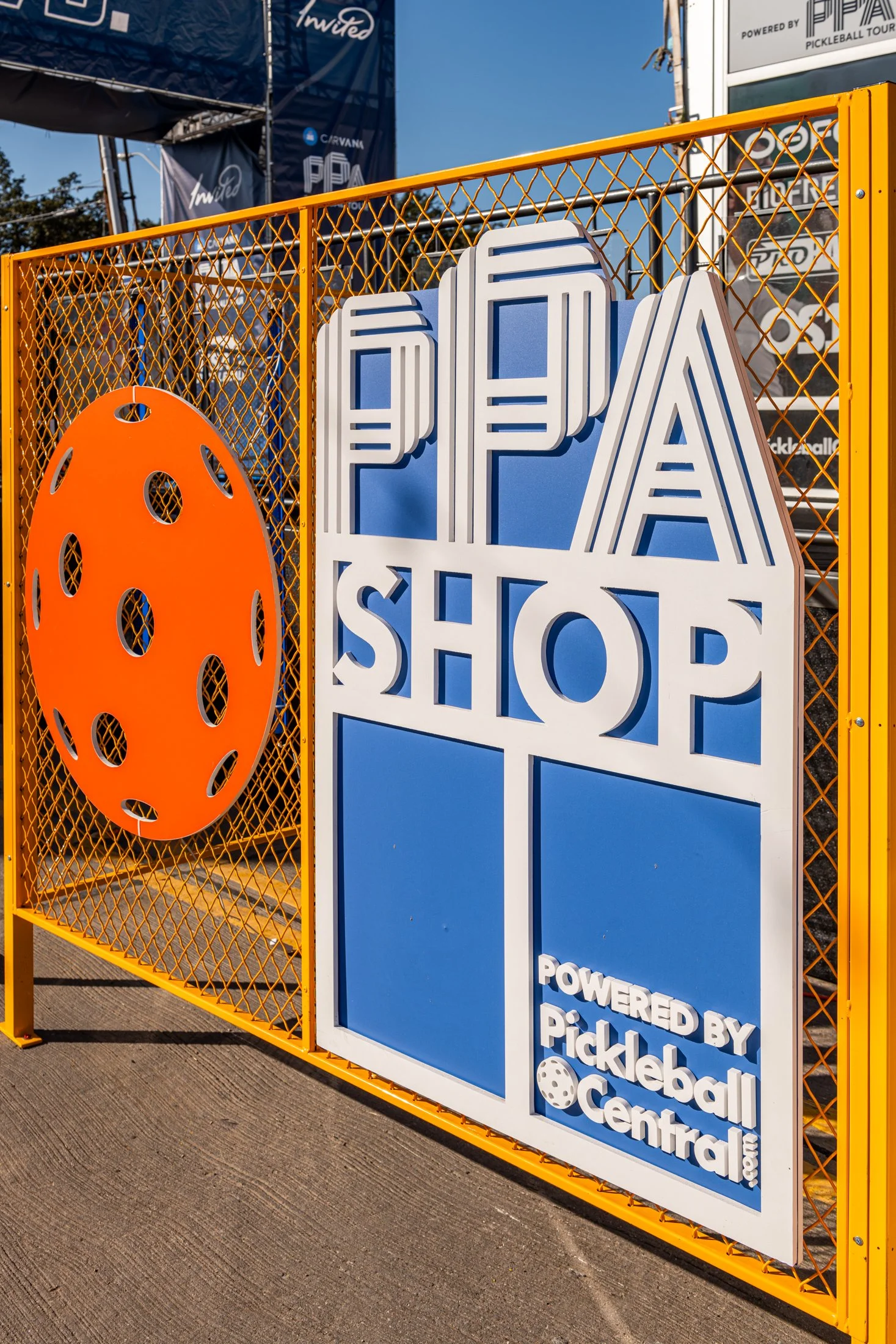 PPA Shop signage on yellow mesh gate featuring an orange pickleball design, promoting pickleball equipment and apparel, powered by Pickleball Central.