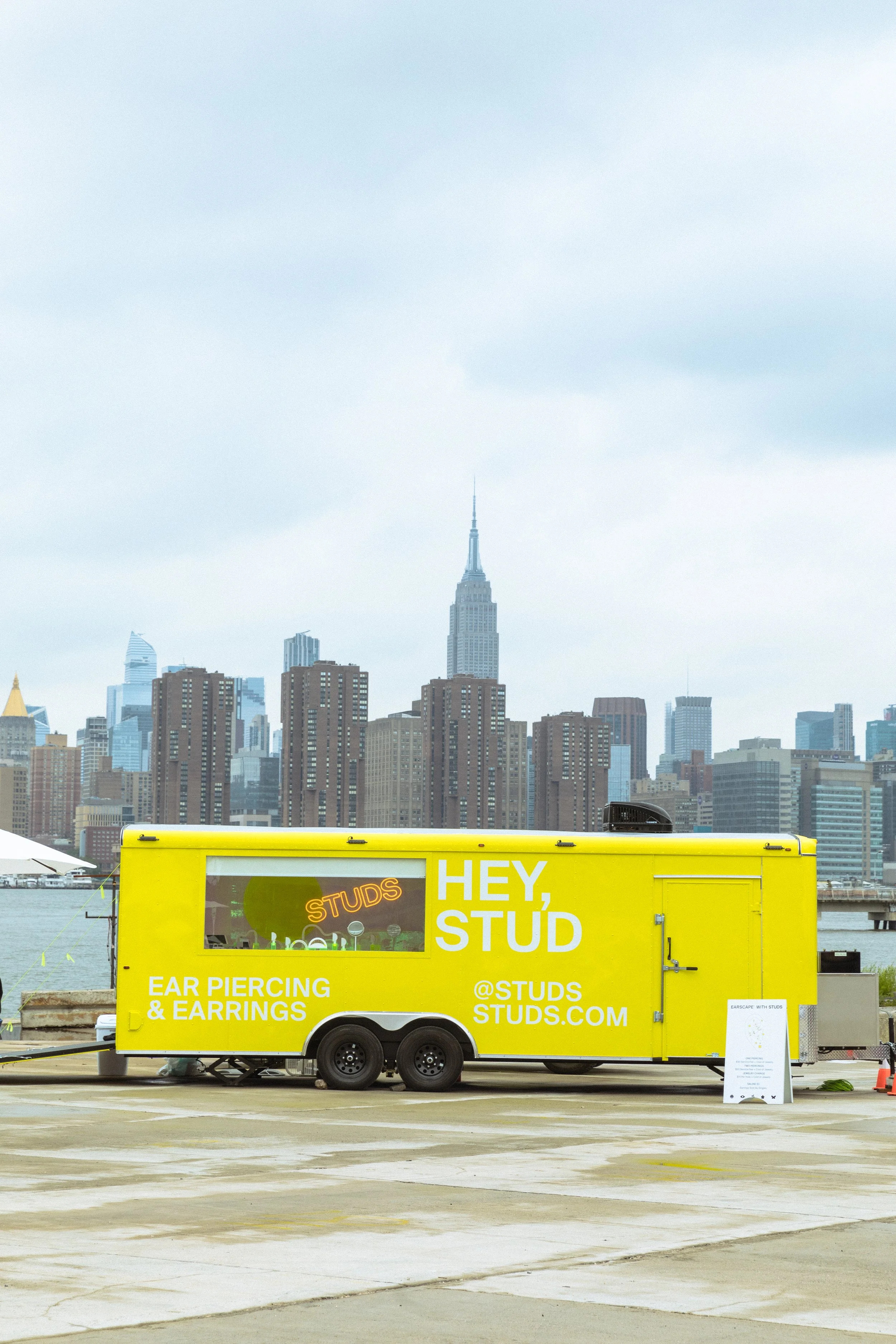 Yellow mobile ear piercing trailer with "HEY, STUD" and "EAR PIERCING & EARRINGS" written on it, parked near a waterfront with a city skyline in the background, including tall buildings.