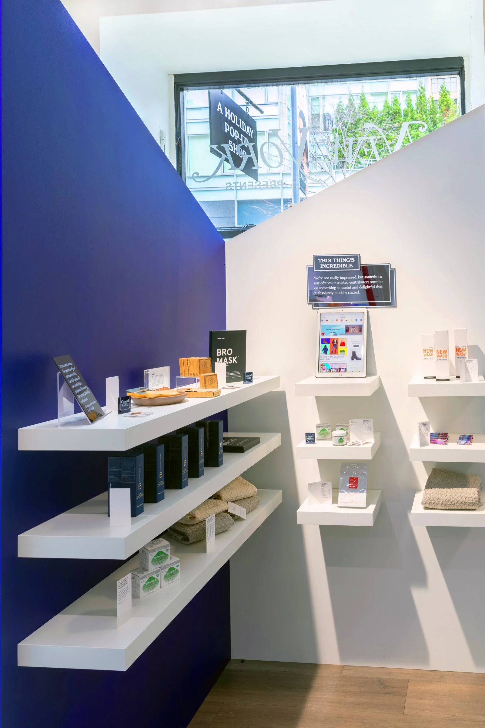 A retail display in a store with white shelves holding skincare products and towels. A sign in the background mentions "A Holiday Pop-In Shop." Blue wall and wooden floor are also visible.