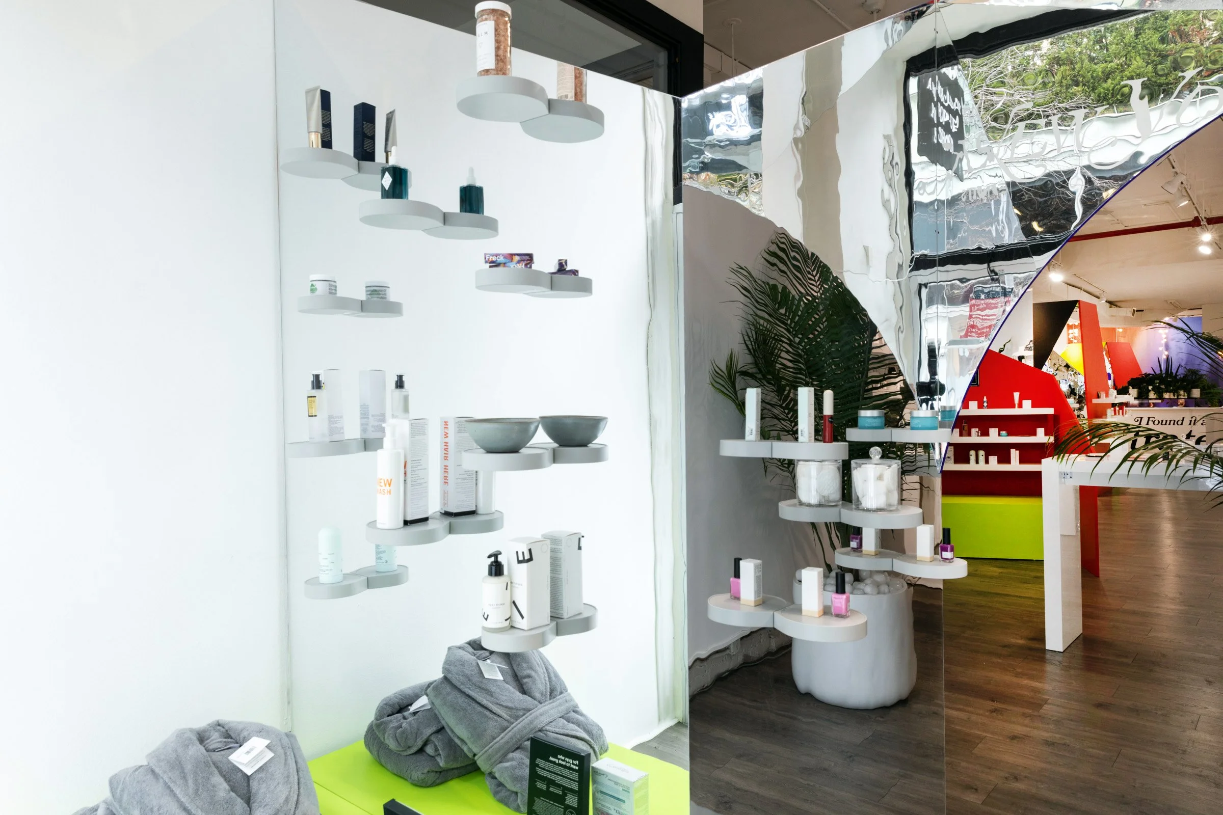 Modern retail display with beauty and skincare products on white floating shelves, set against a mirrored wall. The display features various bottles, jars, and boxes, with some gray towels folded at the bottom. Indoor plants and colorful decor elements are visible in the background.