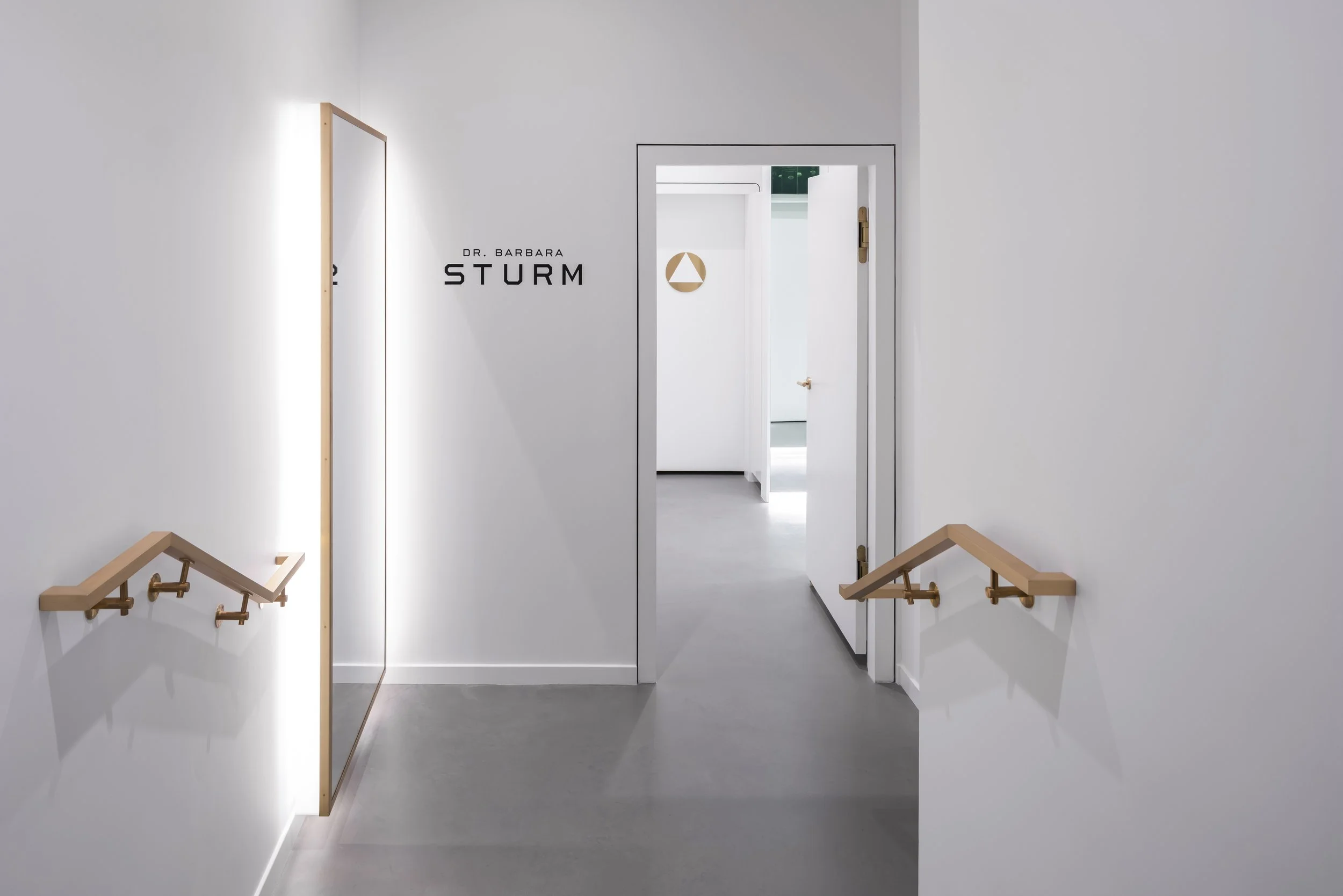 Minimalist hallway with white walls, wooden handrails, and "Dr. Barbara Sturm" signage.