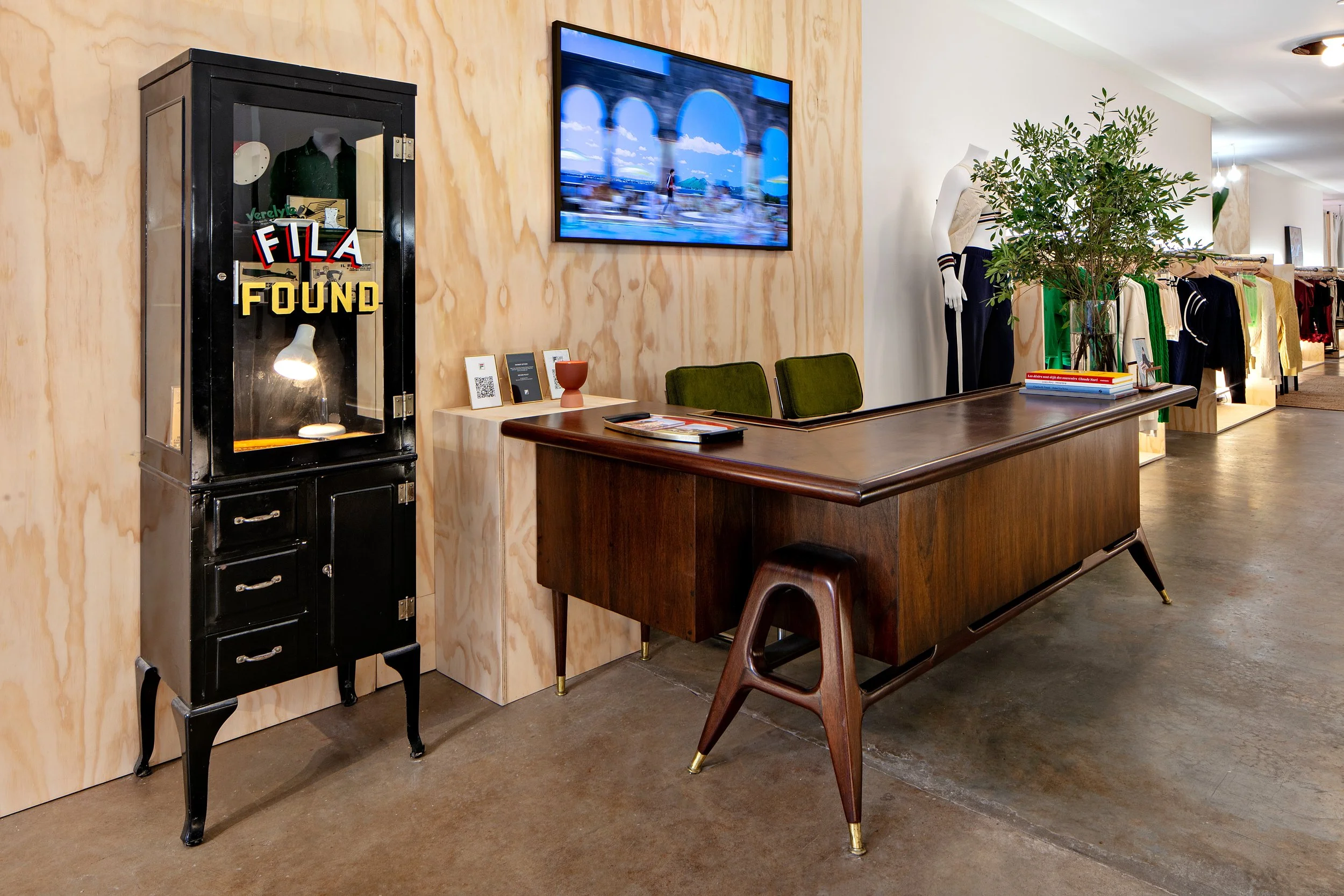 Office interior with a vintage display cabinet labeled 'FILA FOUND,' a wooden desk with green chairs, a TV on the wall, and clothing racks in the background.