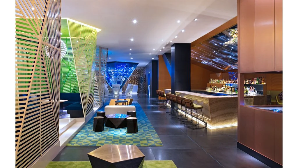 Modern bar interior with geometric design, illuminated bar counter, stools, patterned carpet, and decorative partitions.