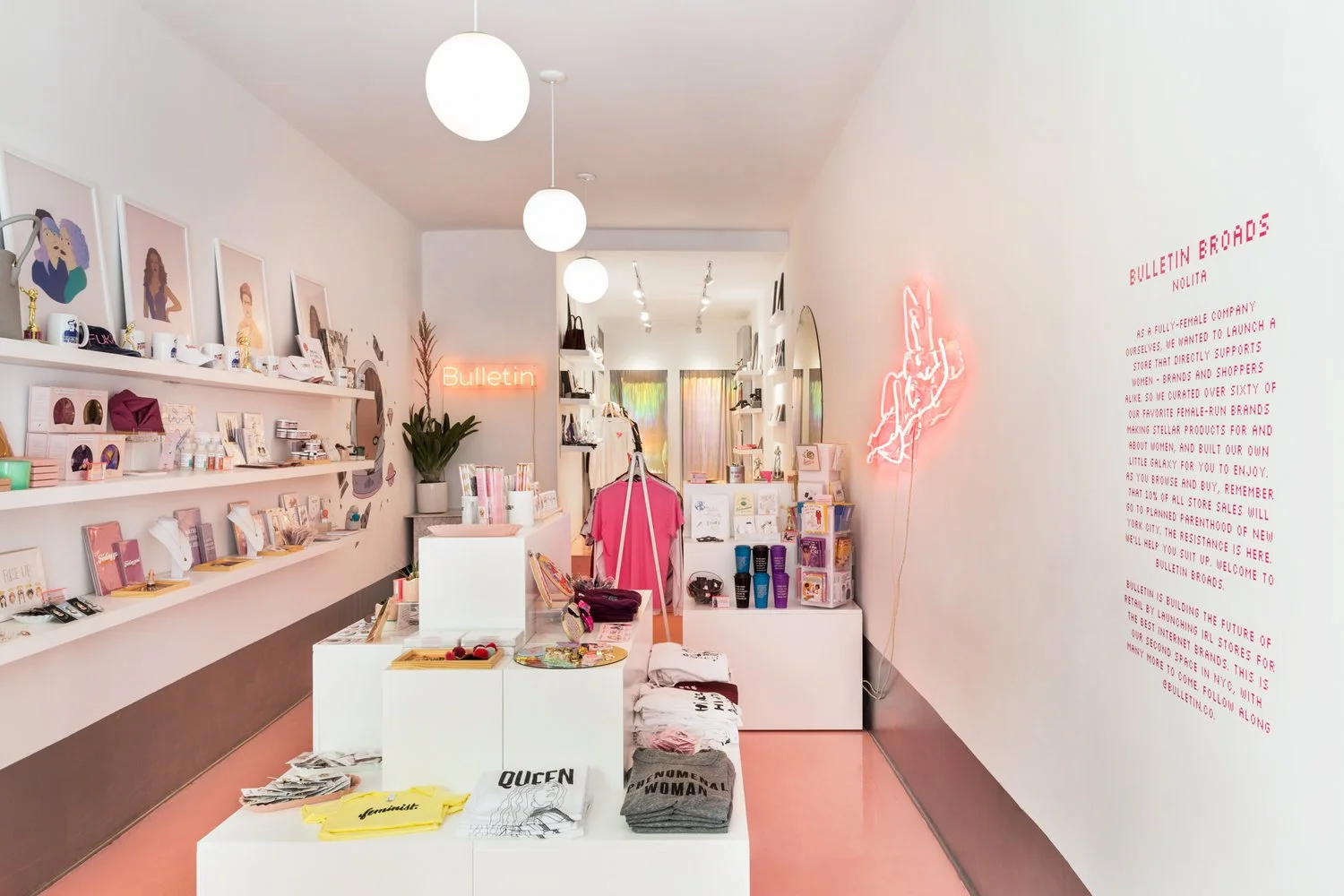 Interior of a boutique shop with white walls and products displayed on shelves, including clothes, mugs, and decor. A neon sign that says 'Bulletin' and a pink neon art piece are on the wall. Text on the right wall explains the store's female empowerment theme.