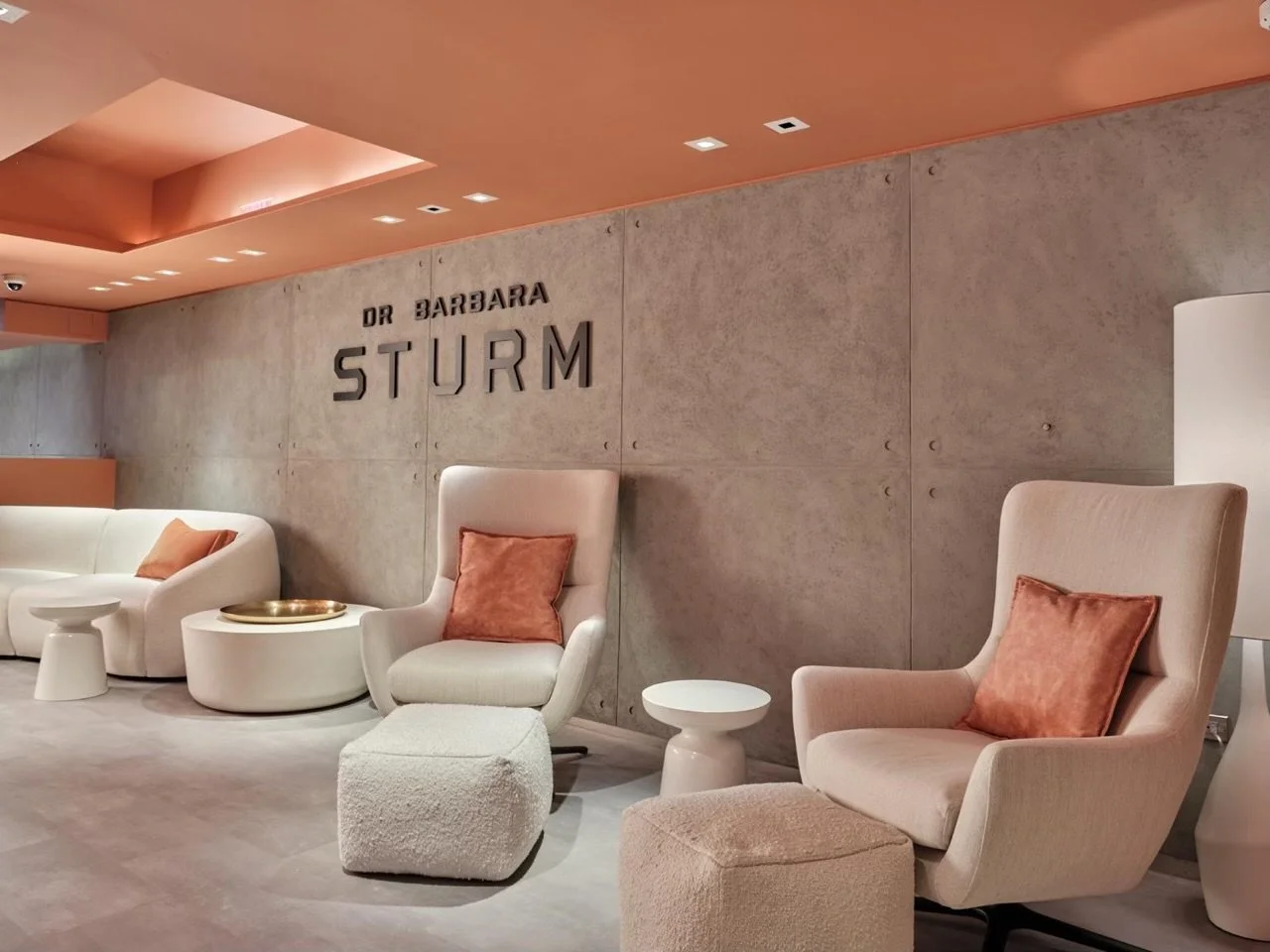 Modern waiting room with beige chairs, a sofa, and orange accents, featuring the text "DR BARBARA STURM" on a concrete wall.