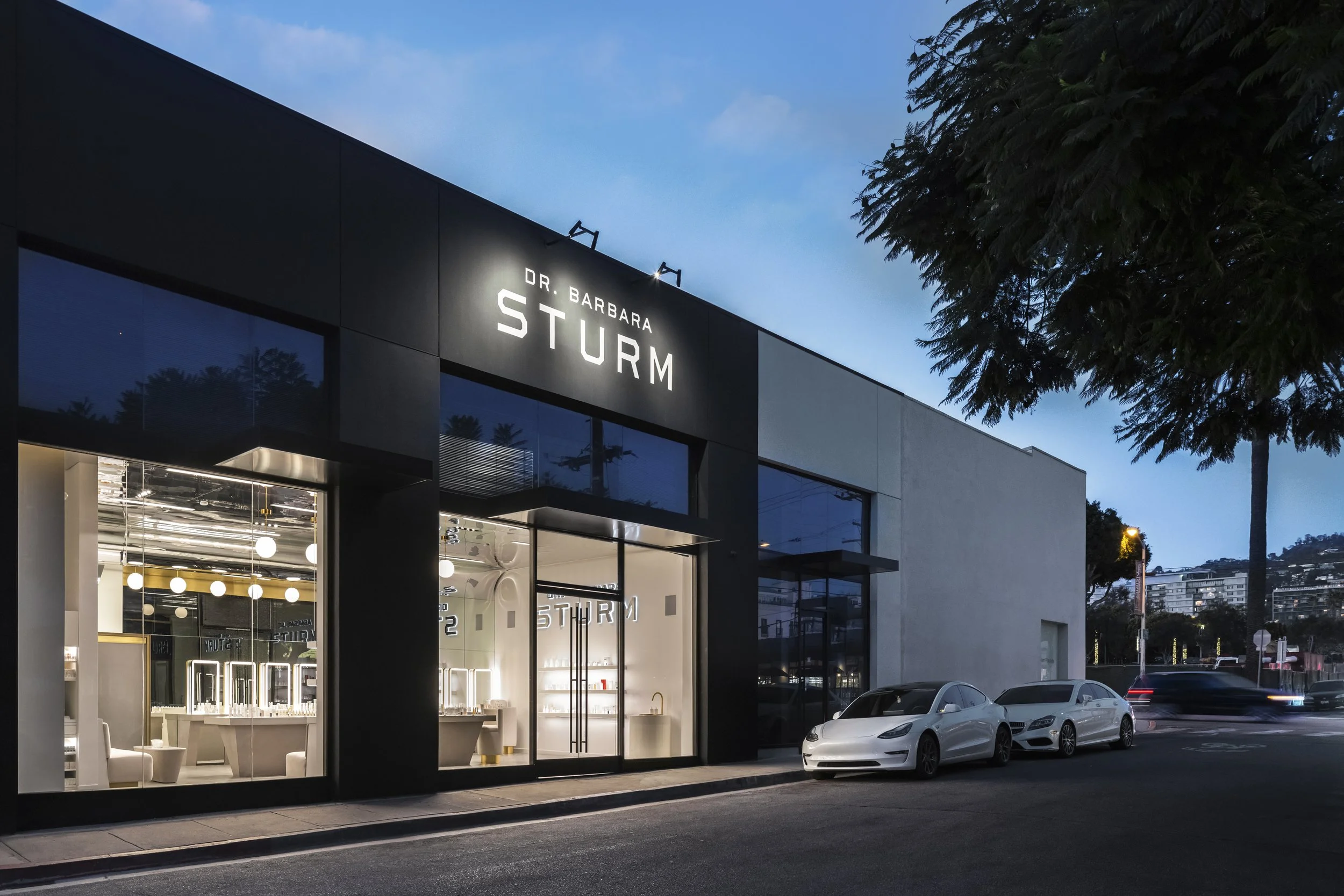 Exterior of the Dr. Barbara Sturm store at dusk, featuring large windows displaying a modern interior with illuminated displays, and two white cars parked outside on the street.