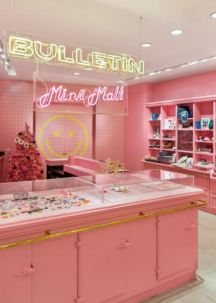 A pink-themed store interior with neon signs reading 'BULLETIN Mini Mall'. A pink Christmas tree with ornaments stands near a wall with a yellow smiley face design. Display cases contain various merchandise.