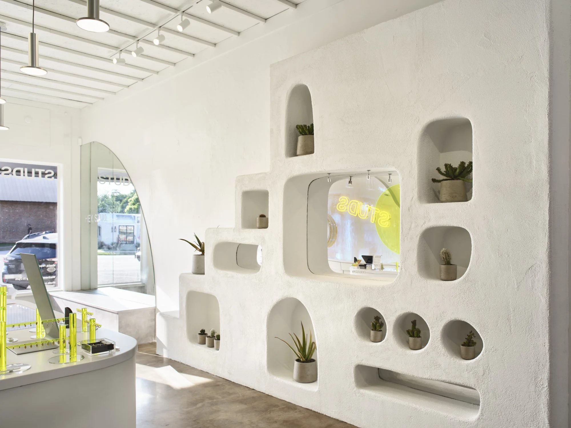A modern, minimalist room with white textured walls featuring built-in shelves displaying small potted plants. The interior has a clean, bright design with track lighting on the ceiling and a large rounded window with views of the outside. A counter with illuminated yellow items is positioned on the left side of the room.