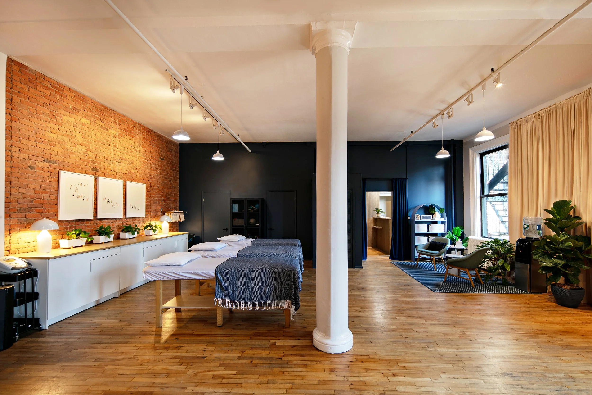 Spacious loft with exposed brick wall, massage tables, white pillar, wooden floors, a seating area, plants, and track lighting.